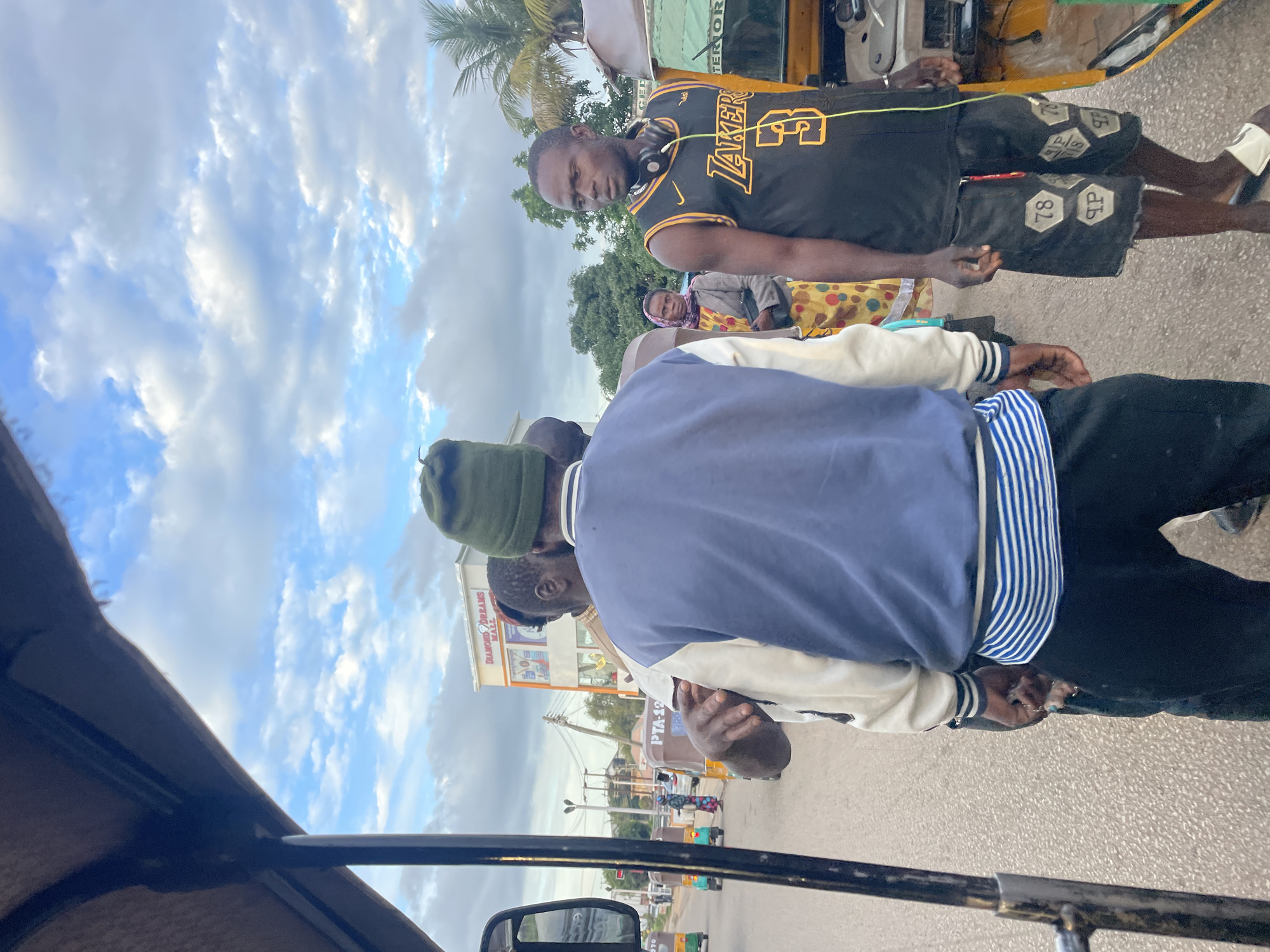 The image shows a street scene in Jos, Nigeria, with several people and vehicles. In the foreground, a man wearing a blue and white jacket and a green beanie is facing away from the camera, talking to another man. The second man is wearing a black basketball jersey with "LAKERS" and the number "3" on it, along with black shorts and headphones around his neck. Behind them, a woman in a colorful patterned dress and a grey jacket is looking at the camera. Several auto-rickshaws, known as "Keke Napeps" in Nigeria, are also visible on the road, along with a building in the background labeled "DIAMOND DREAMS MALL." The sky is partly cloudy.