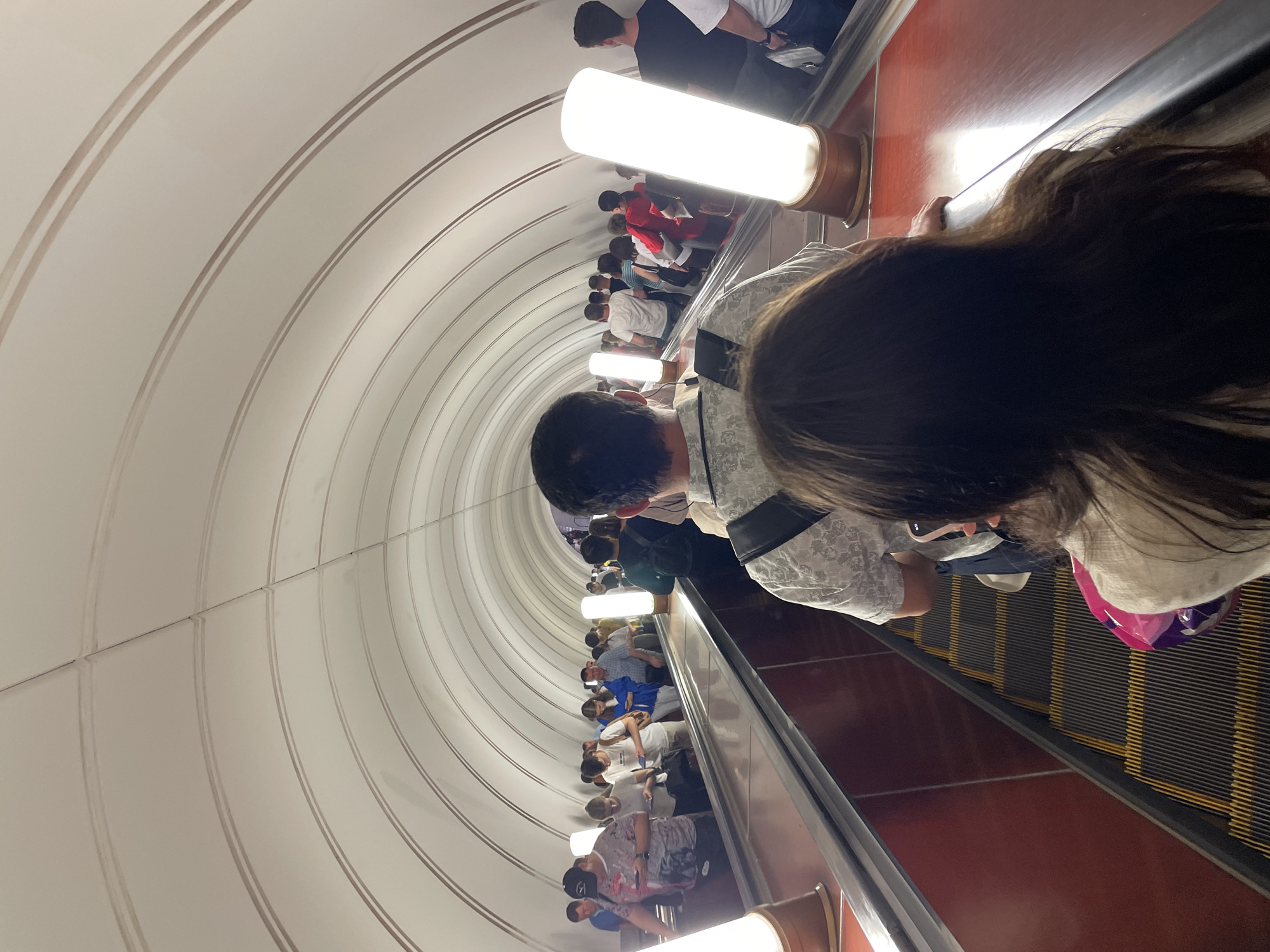 The image captures a scene inside the Moscow Metro system in Russia. People are descending on a long escalator. The tunnel is lined with white tiled arches. Artificial light fixtures illuminate the tunnel. The focus of the photograph is on the individuals traveling in front of the photographer. Other passengers can be seen descending behind them. The escalator steps are dark gray with yellow lines. The sides of the escalator are red.