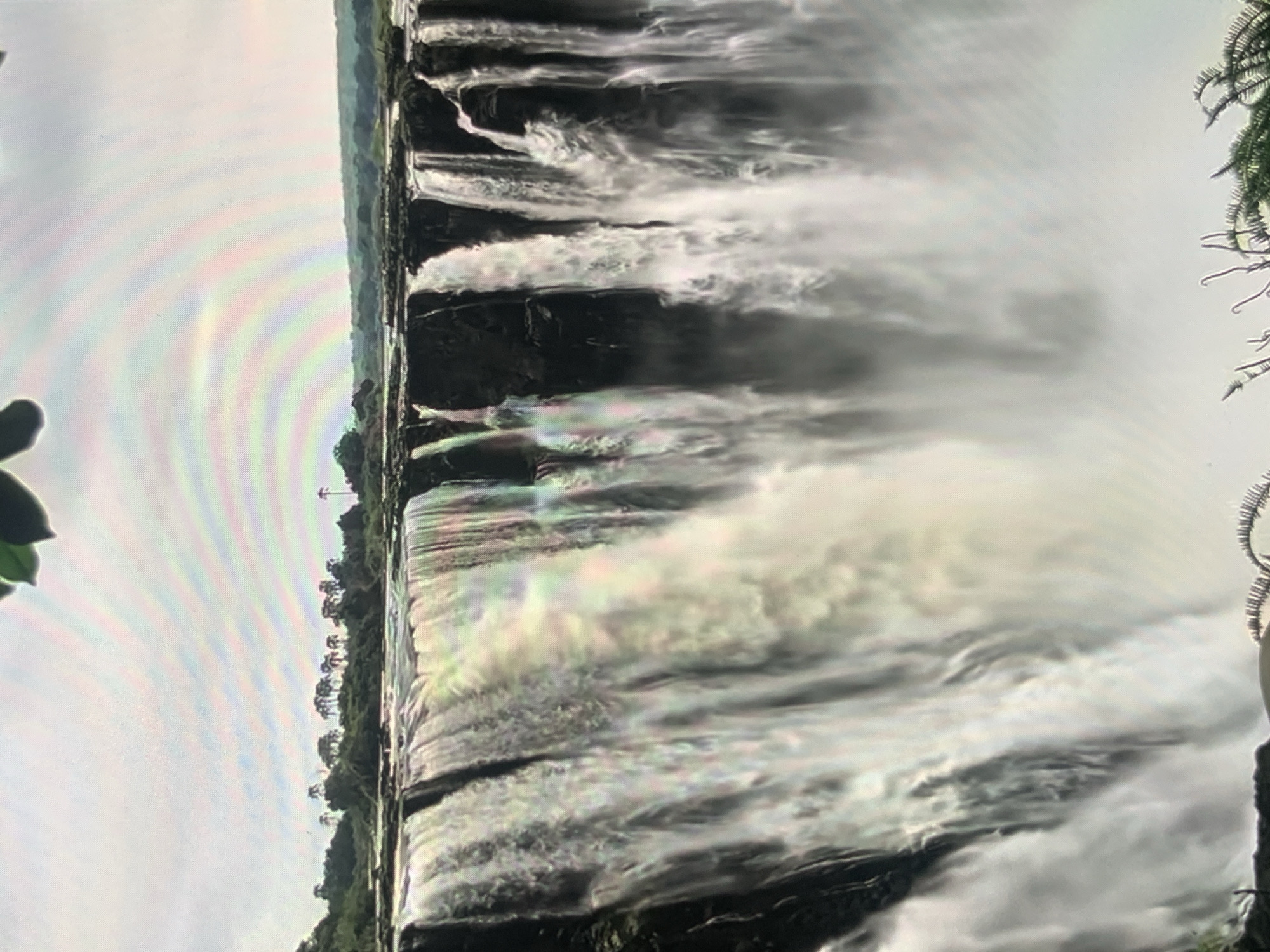 The image shows a wide waterfall cascading over a cliff face. The water is white and frothy, with spray rising from the base. In the background, there is a line of trees and a hazy sky with a faint rainbow. The foreground shows the edge of the waterfall and some vegetation. There are no people visible in the image. The location is not Stavropol, Russia; this is a natural landscape.