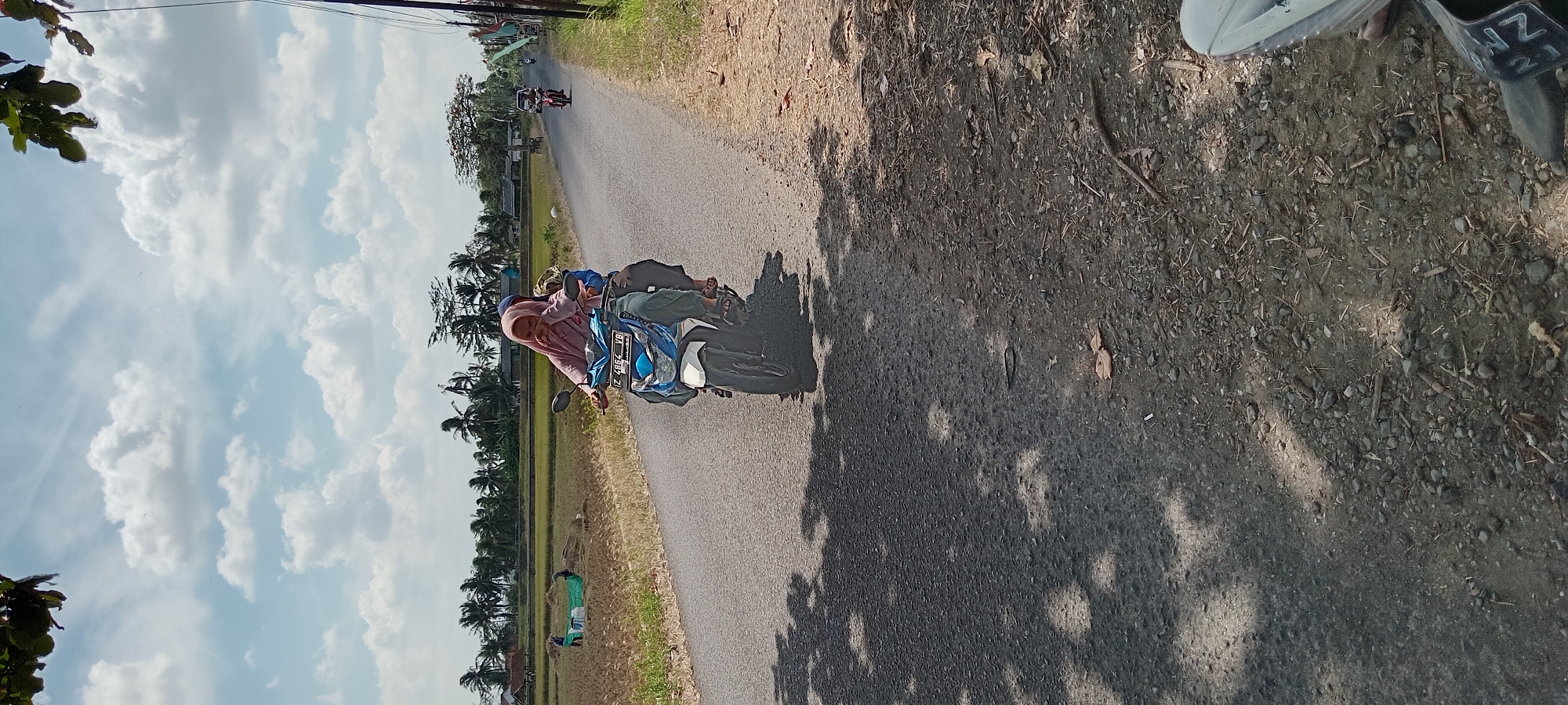 The image depicts a sunny day with scattered clouds in Karangpawitan, Indonesia. The foreground is dominated by a paved road with gravel and debris on its shoulder. Sunlight casts dappled shadows from overhead foliage onto the road.
In the midground, a blue and white scooter is traveling away from the viewer. A woman wearing a pink hijab and a dark blue headscarf is driving the scooter. She is looking downwards and to her left. A child, also wearing a dark head covering, is seated behind her on the scooter. The scooter has a license plate that reads "F Z 4864 VB".
Beyond the road, a rural landscape unfolds. A field of dry rice stalks is visible to the left, with two individuals in the field working with a large green tarp. In the background, a line of palm trees and other vegetation marks the horizon, with a few buildings scattered amongst them. Another scooter is visible further down the road, also moving away from the viewer. A utility pole with wires stands to the right of the road. The overall scene suggests a typical day in a rural Indonesian setting.