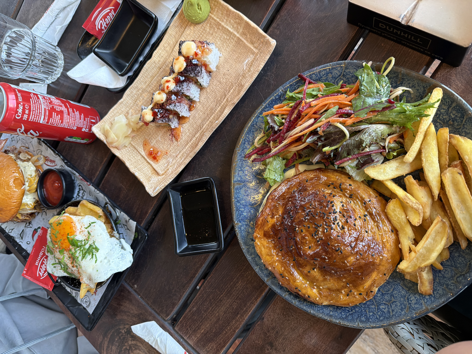 The image shows a table at an outdoor restaurant setting. The table, made of dark wooden planks, is laden with several plates of food and accompanying items. A plate with what appears to be sushi rolls, garnished with sauces and a small dollop of green wasabi, occupies the top-center portion. To the right, on a blue plate, is a round, golden-brown savory pie topped with sesame seeds. Alongside it are french fries and a mixed salad. To the left, a burger with a fried egg is visible on a smaller black tray. A can of Coca-Cola is partially visible in the top-left quadrant. Black square dishes filled with dark sauces are scattered among the plates. The brand "Happy" is printed on some of the packaging, along with "DUNHILL" on a box placed near the blue plate. The scene appears to be in Varna, Bulgaria, based on the Cyrillic text.