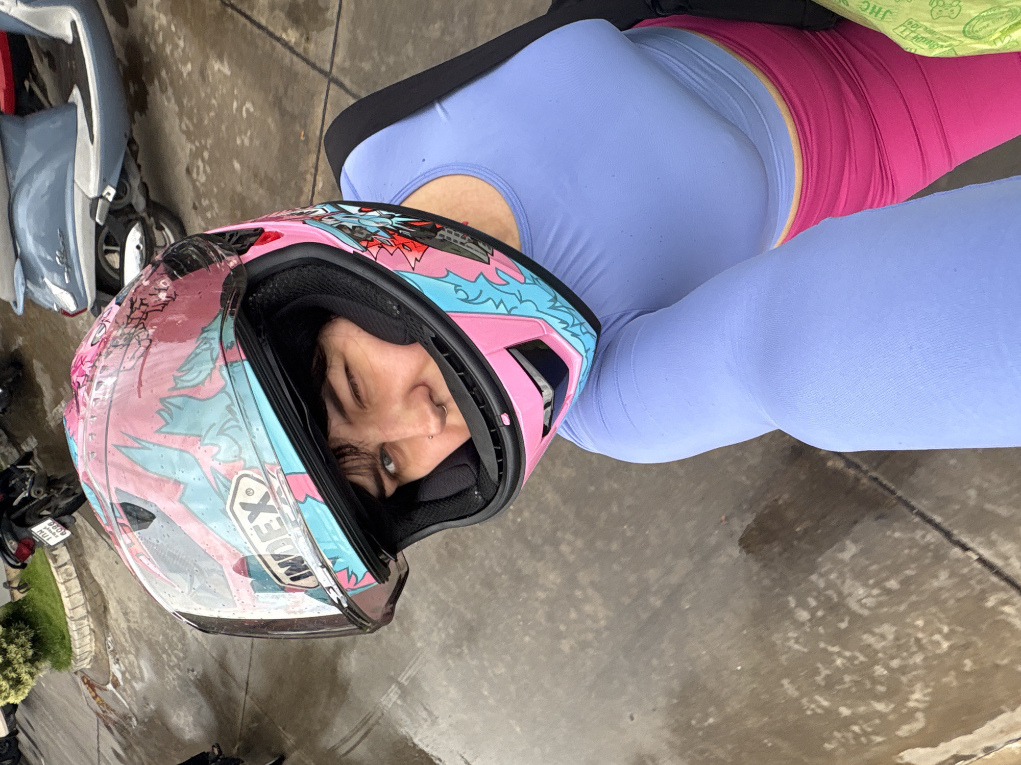 A person wearing a pink and blue motorcycle helmet and a light blue long-sleeved shirt is taking a selfie. The person has dark hair and is winking, with a nose ring and an earring visible. They are wearing bright pink pants. In the background, there are several parked scooters and motorcycles, and the ground is wet, suggesting it has rained recently. The scene appears to be an outdoor parking area, possibly in the city of Nong Pa Khrang, Thailand.