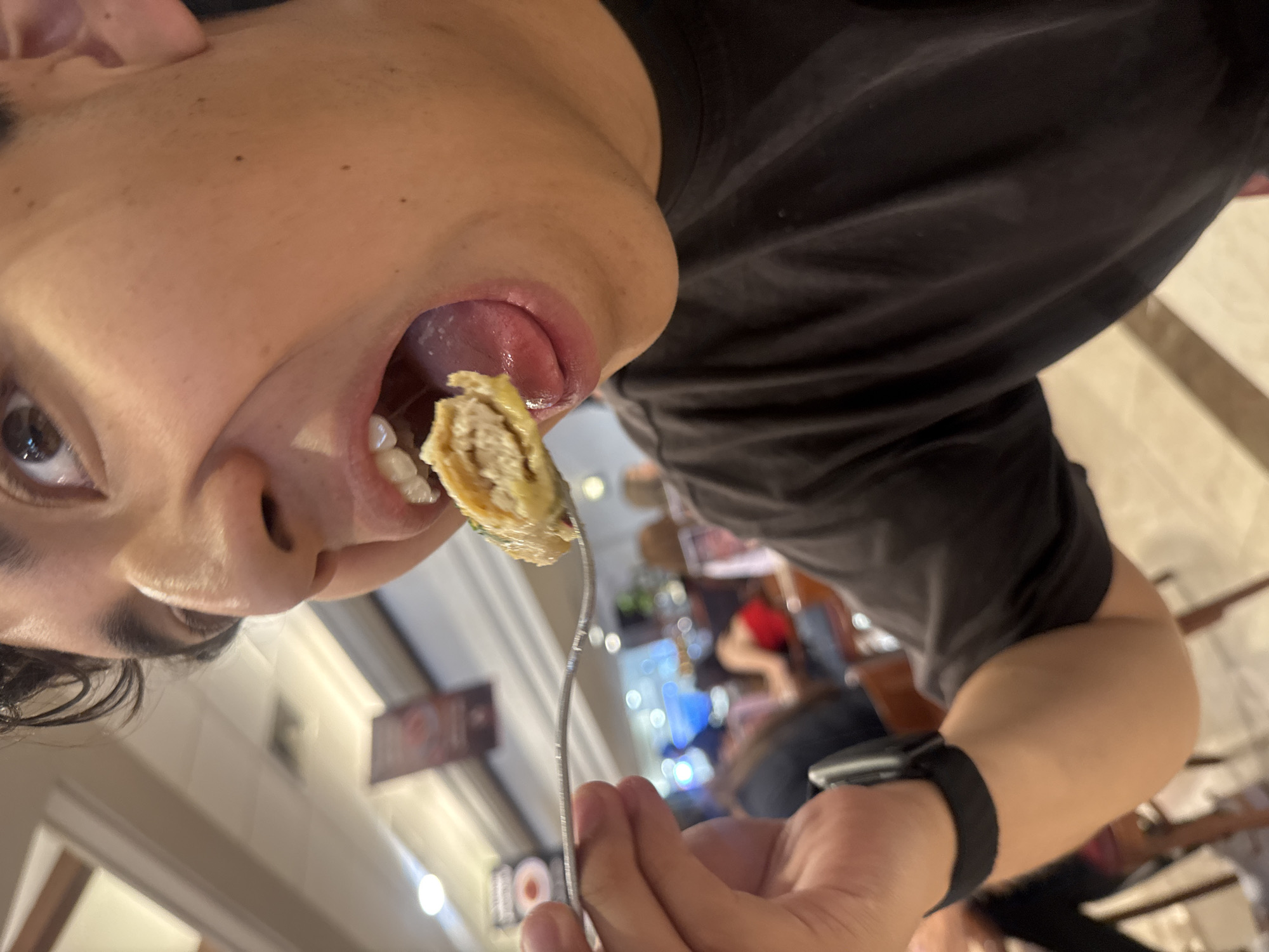 A person is shown close up, head tilted back and to the left, about to eat a roll of food on a fork. The person's mouth is open with their tongue extended. The person is wearing a black shirt and a black wristband, possibly a smartwatch. In the blurred background, other people are seated at tables in what appears to be a restaurant. Based on signage visible and common knowledge, this image appears to have been taken in the city of Puebla, Mexico.