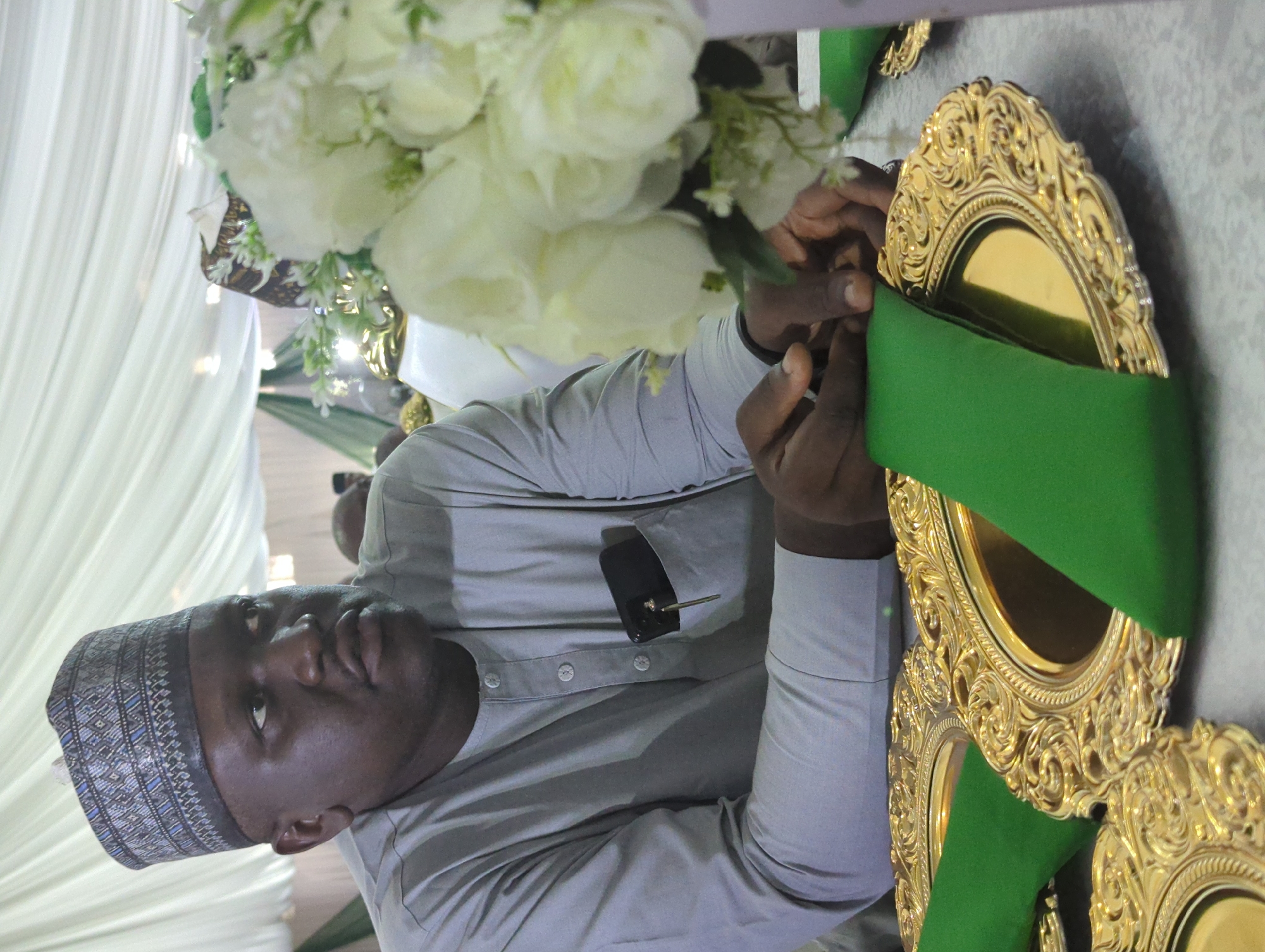 The image depicts a medium shot of a man wearing a grey traditional outfit and a patterned cap. He is seated at a table adorned with golden ornate plates and green napkins. A bouquet of white flowers is visible to his right. A mobile phone and a key are tucked into his chest pocket. His hands are resting on the table in front of him. The background consists of draped white fabric, suggesting an indoor event setting, possibly in Unguwar Rimi, Nigeria.