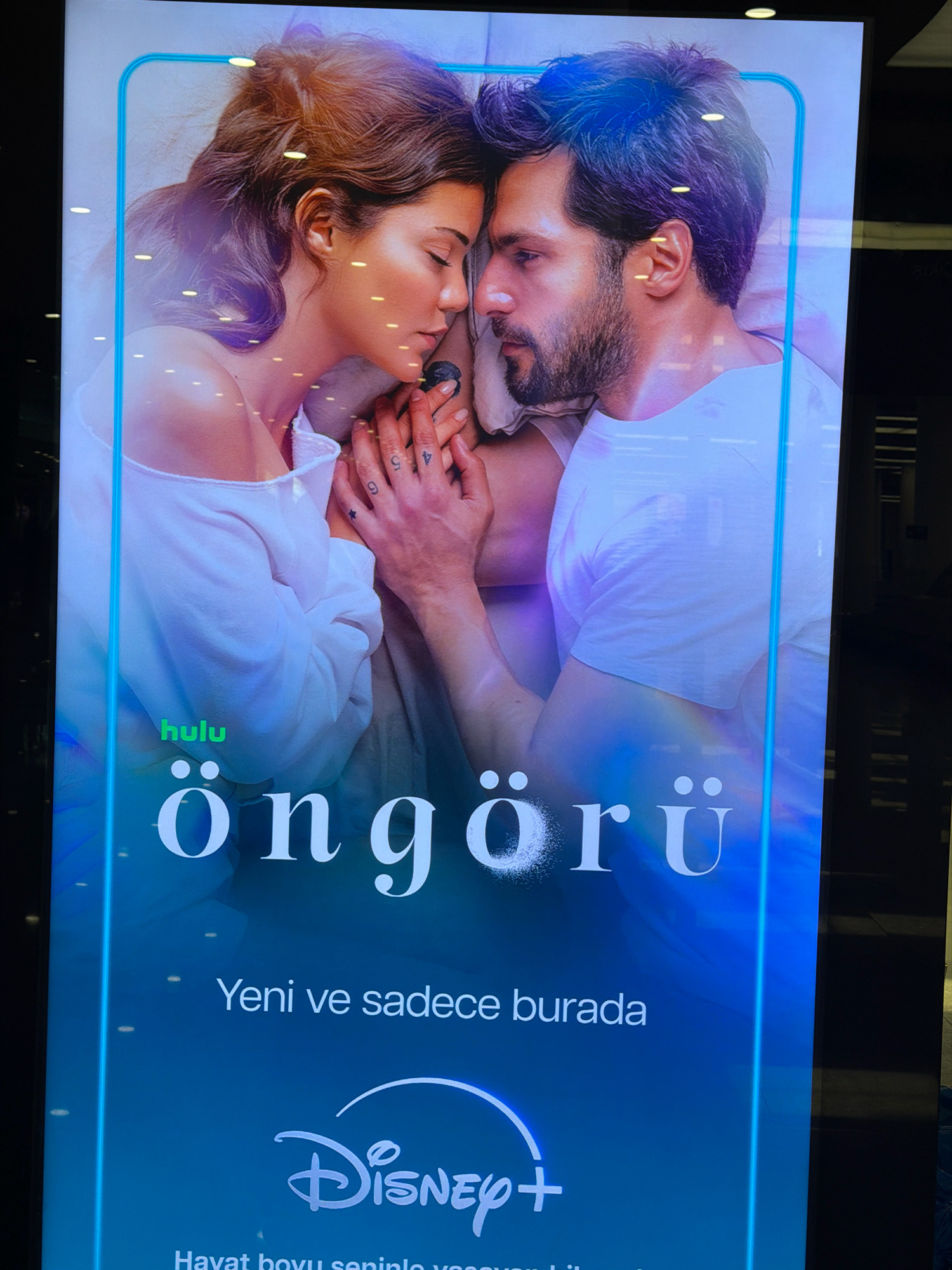 A vertical poster displays a close-up, intimate image of a man and a woman lying down, their heads touching. The woman is on the left, her eyes closed, and the man is on the right, also looking down with his eyes closed. Their hands are intertwined. Both are wearing casual, light-colored tops. The poster features text in Turkish, including "hulu," "öngörü," "Yeni ve sadece burada," and the Disney+ logo. The overall color scheme is a blend of soft blues and purples, creating a serene and romantic mood. The poster is illuminated, with some reflections on its surface, suggesting it is on display in a public place, possibly a shop window or an advertisement screen. The context of Gaziemir, Turkey, implies this poster is located within a commercial or public area in that city.