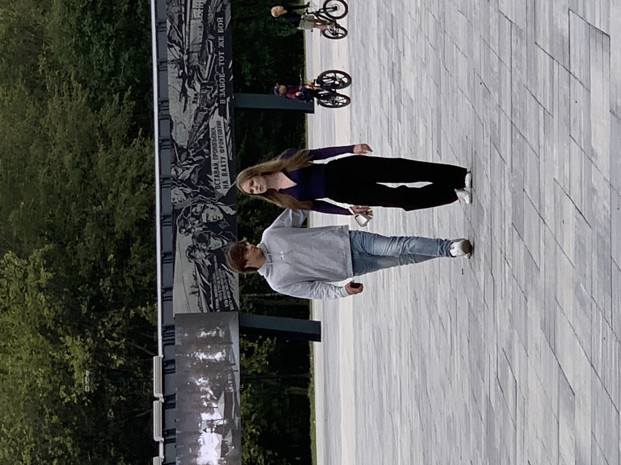 In Prokopyevsk, Russia, two young adults, a male and a female, walk together on a paved plaza. The male, wearing a grey hooded sweatshirt and blue jeans, has his right arm around the female's shoulders. The female, dressed in a dark long-sleeved top and black pants, walks to his left. Both are looking forward. In the background, a large mural depicting historical scenes and text is visible on a wall, bordered by trees. Two children are riding bicycles further down the plaza. The ground is paved with rectangular gray stones.