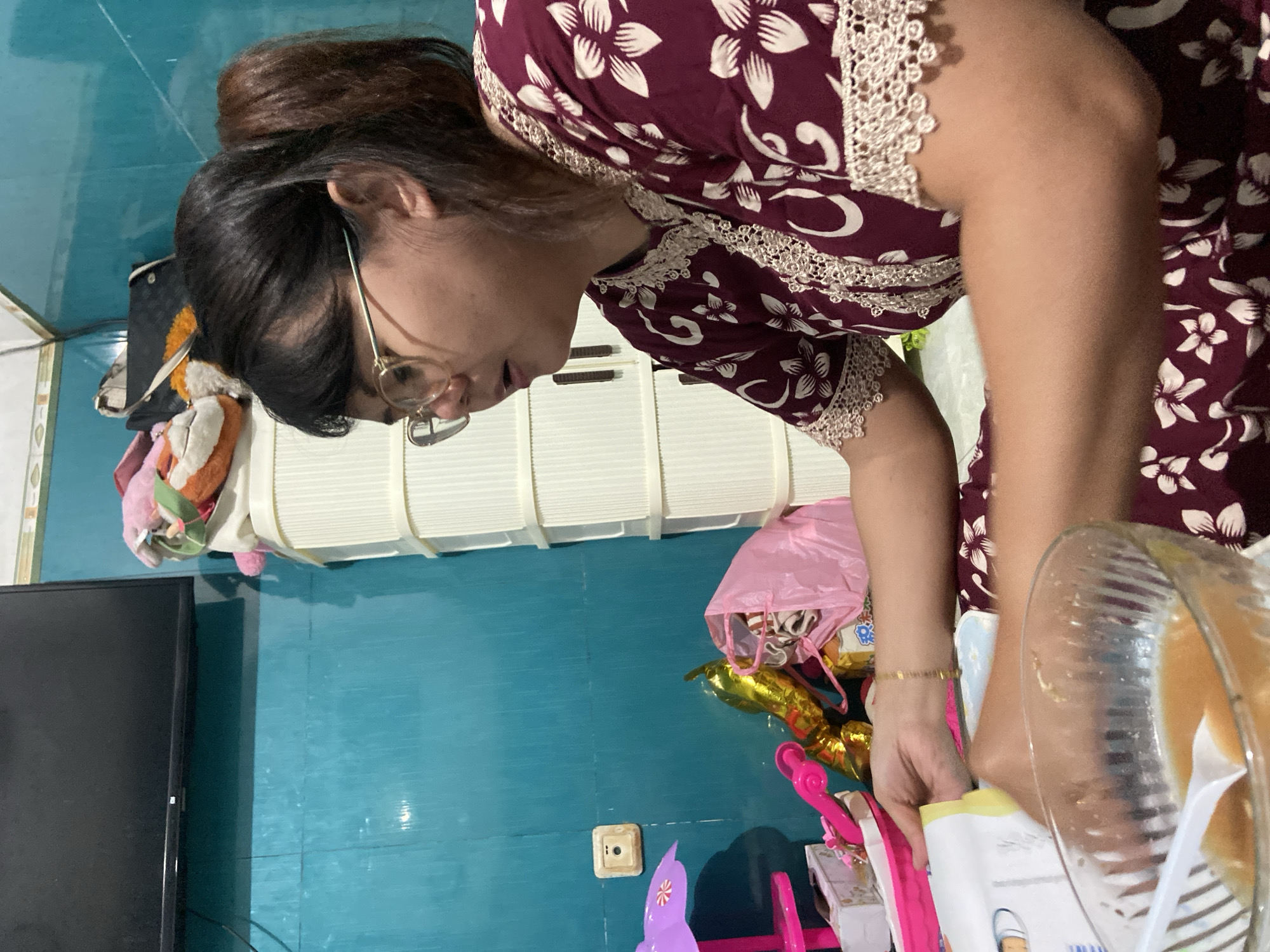 A person wearing glasses and a floral patterned dress is reaching into a pile of items that include a book, a pink bag, and gold-colored balloons. The person is located in a room with a blue wall and a black television set. The location is possibly in Gelam, Indonesia.