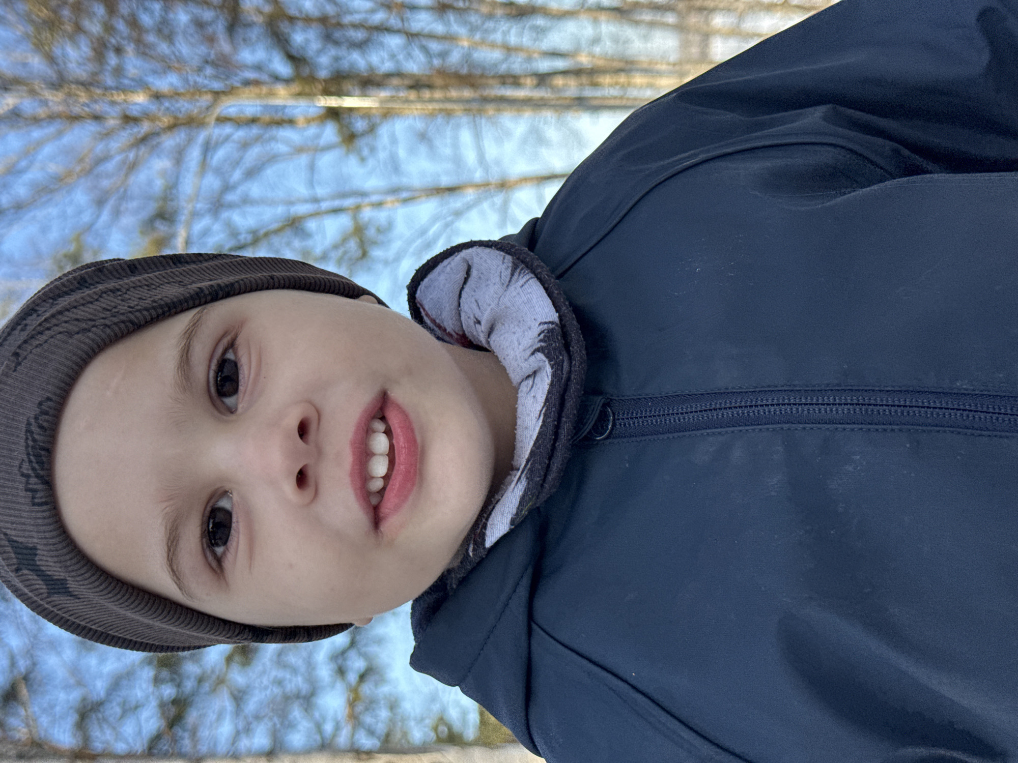 A young child, possibly male, is depicted from a low-angle perspective. The child is wearing a dark-colored beanie and a dark blue jacket with a lighter-colored lining visible around the neck. The child's face is slightly tilted and they are looking directly at the camera with their mouth slightly open. The background appears to be a blurred outdoor scene, likely trees and sky, suggesting a natural environment. There is no indication of the location being Chaikovsky, Russia, or any urban setting. The image primarily focuses on the child and their attire against a natural backdrop.