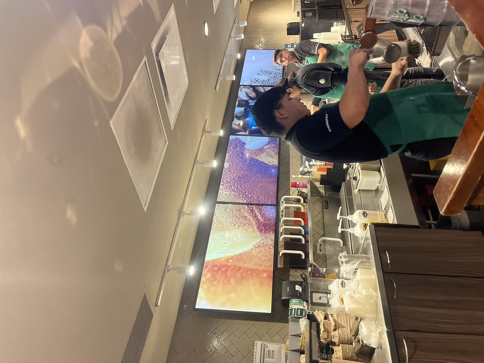 A photograph taken inside a Starbucks in Mexico City, Mexico shows two people working behind the counter. One individual is using a coffee machine to prepare a drink. A second person is visible in the background. The interior has multiple large displays showing abstract visuals. The counter area includes various dispensers, containers, and supplies necessary for drink preparation. The location appears to be well-lit, with ceiling-mounted lighting fixtures.