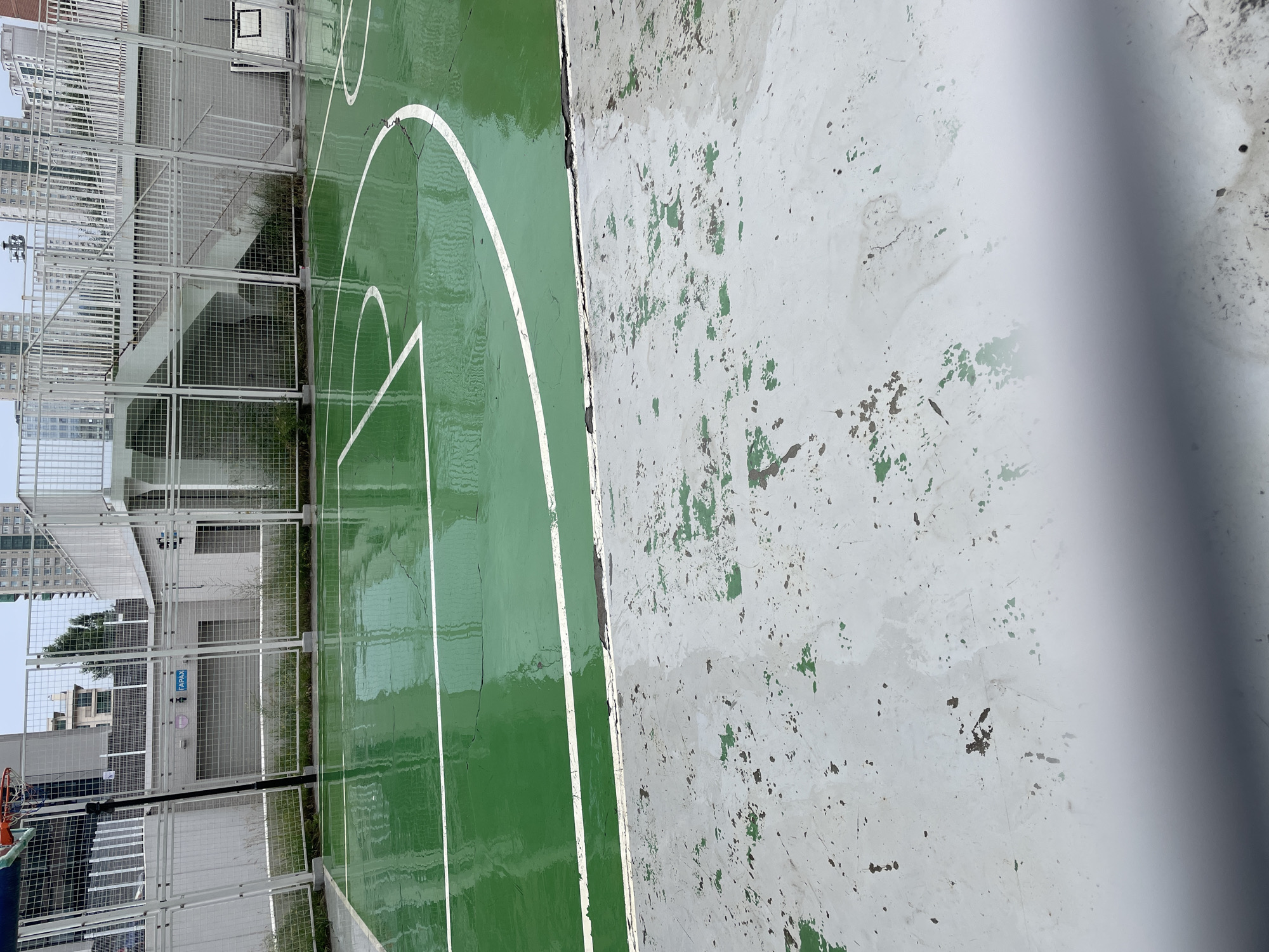 The image shows a wet outdoor basketball court with green painted surface marked with white lines. The court is bordered by a white metal fence and a concrete area with peeling paint. In the background, there are multi-story buildings and a staircase with white railings. A basketball hoop is partially visible on the left. The sky is overcast.