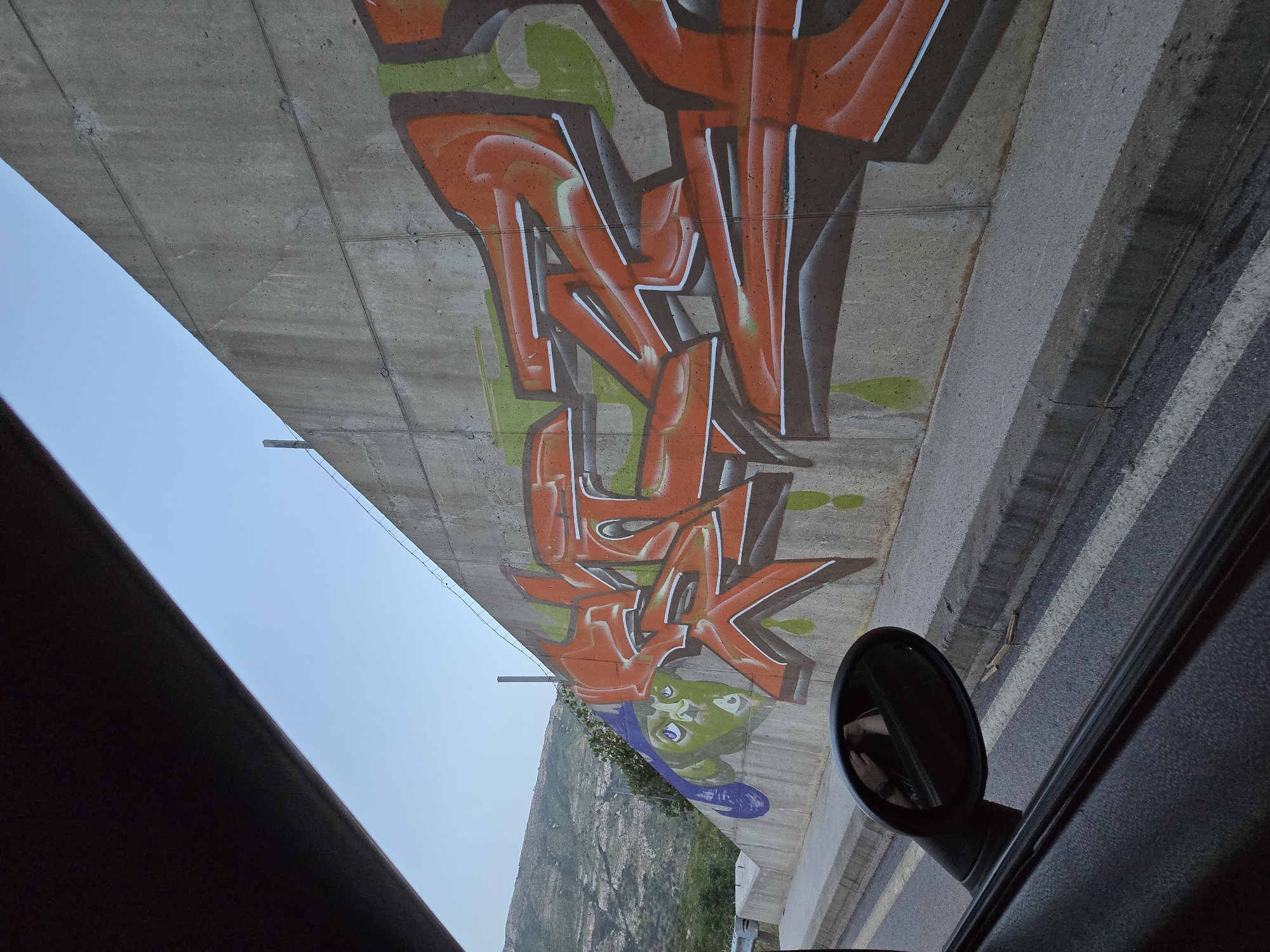 A wide shot, captured from the interior of a vehicle, shows a concrete wall adorned with graffiti. The graffiti features large, orange, stylized lettering with white and dark brown outlines, accented by green splashes. To the left of the lettering, a green, cartoon-like face with purple accents is painted. The concrete wall is part of an elevated structure, as evidenced by its angled orientation and the presence of wires along its top edge. In the background, a mountainous landscape with sparse vegetation is visible under a clear sky. A car's side mirror is reflected in the foreground, showing a partial reflection of the vehicle's interior and a person holding a phone. The scene appears to be a roadside location, indicated by the presence of a curb and road markings. The image is identified as being taken in Klopot, Montenegro.