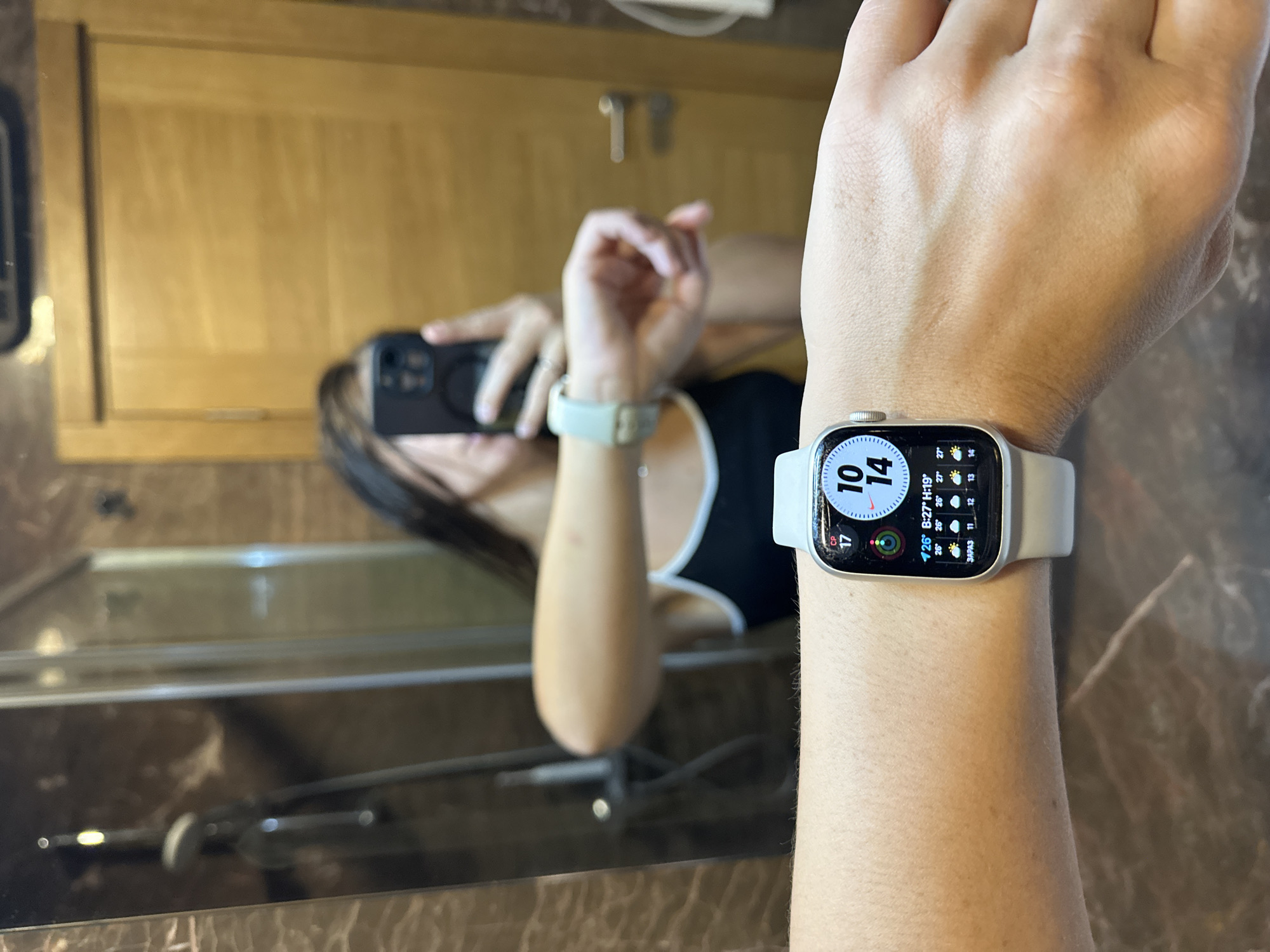 The image captures a person's wrist, adorned with a white Apple Watch, reflecting a mirror in a bathroom. The watch face displays the time as 10:14 and includes weather information, showing a temperature of 26 degrees Celsius, a low of 19 degrees Celsius, and a high of 27 degrees Celsius. The background reveals a mirror reflecting a woman holding a smartphone, her arm extended as she presumably takes a selfie. The bathroom environment features tiled walls with a brown, marble-like pattern, a shower head visible in the reflection, and a wooden door. The overall scene suggests a personal moment captured in a domestic setting. The location is identified as Donji Pobori, Montenegro.