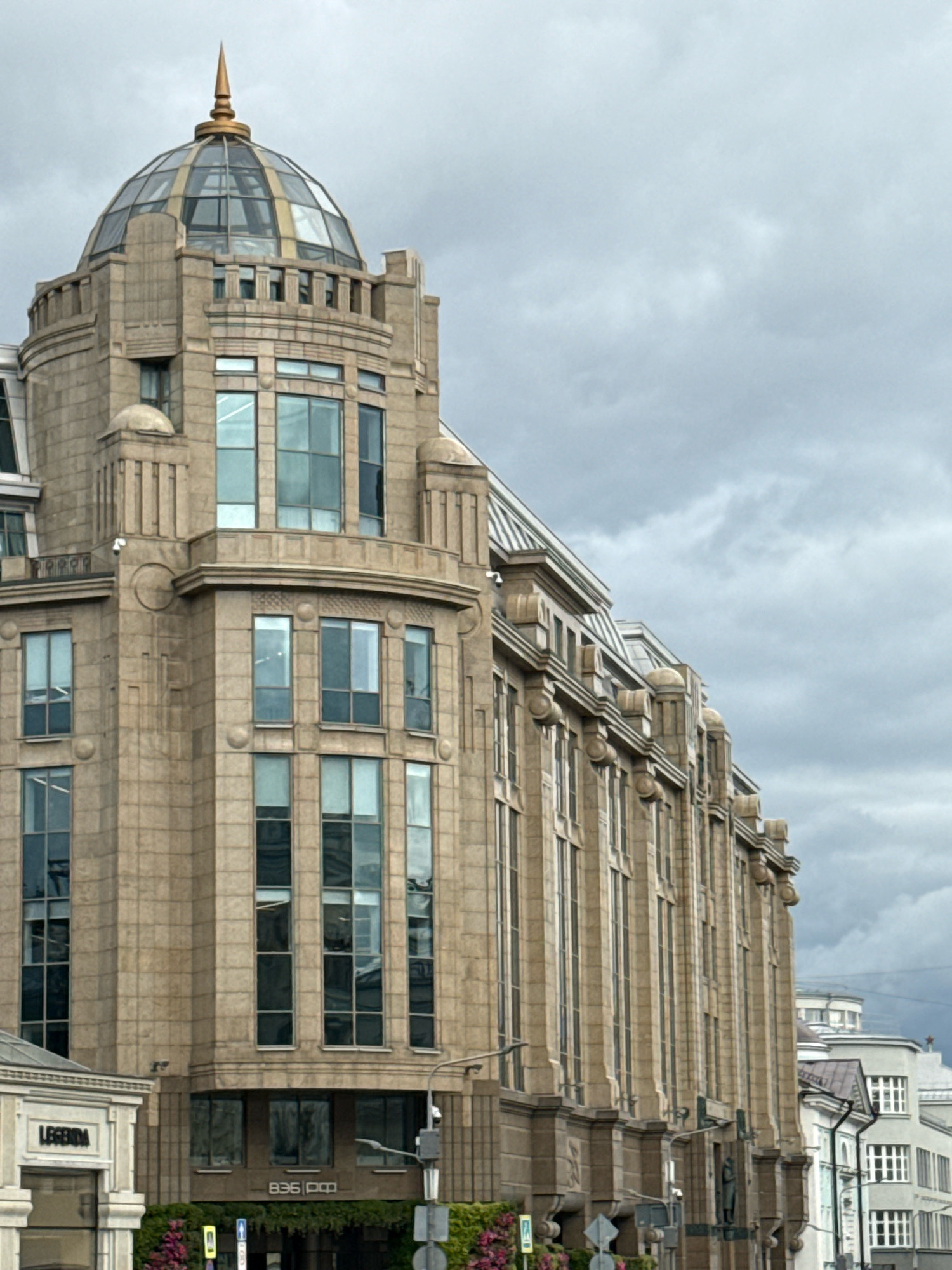 The image shows the exterior of the Central Children's Store on Lubyanka Square in Moscow, Russia. The building is an imposing structure with a distinctive dome topped by a golden spire. The facade is made of light-colored stone blocks and features numerous tall, rectangular windows. Ornate architectural details, such as pilasters and decorative moldings, are visible along the sides of the building. The sky is overcast with grey clouds. In the foreground, a portion of a building with the name "LEGGINDA" is visible, along with some greenery and street signs. The image is taken from a low angle, emphasizing the height and grandeur of the Central Children's Store.