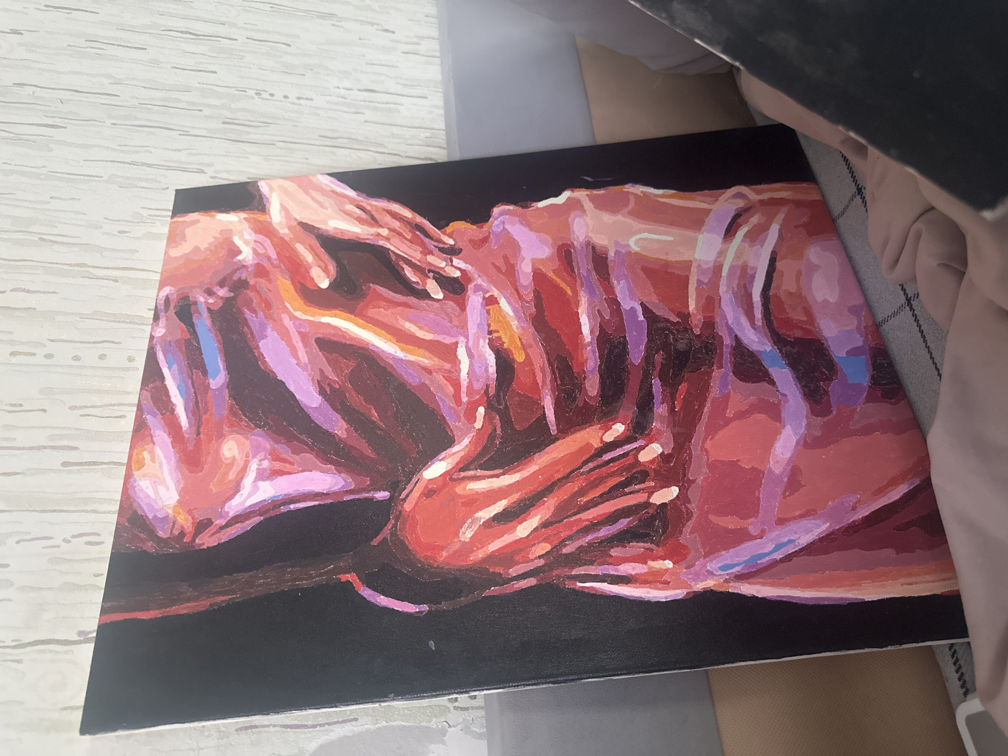 A painting on a canvas sits on a surface, likely a table or desk. The painting features an abstract rendering of two figures, primarily rendered in shades of red, pink, purple, and orange, against a black background. The figures' forms are not clearly defined but seem to depict bodies with visible hands. The surface underneath the painting appears to be textured and is partially obscured by other objects, including a dark-colored object and fabric. The location Высокий, Russia is not visually evident in the image.