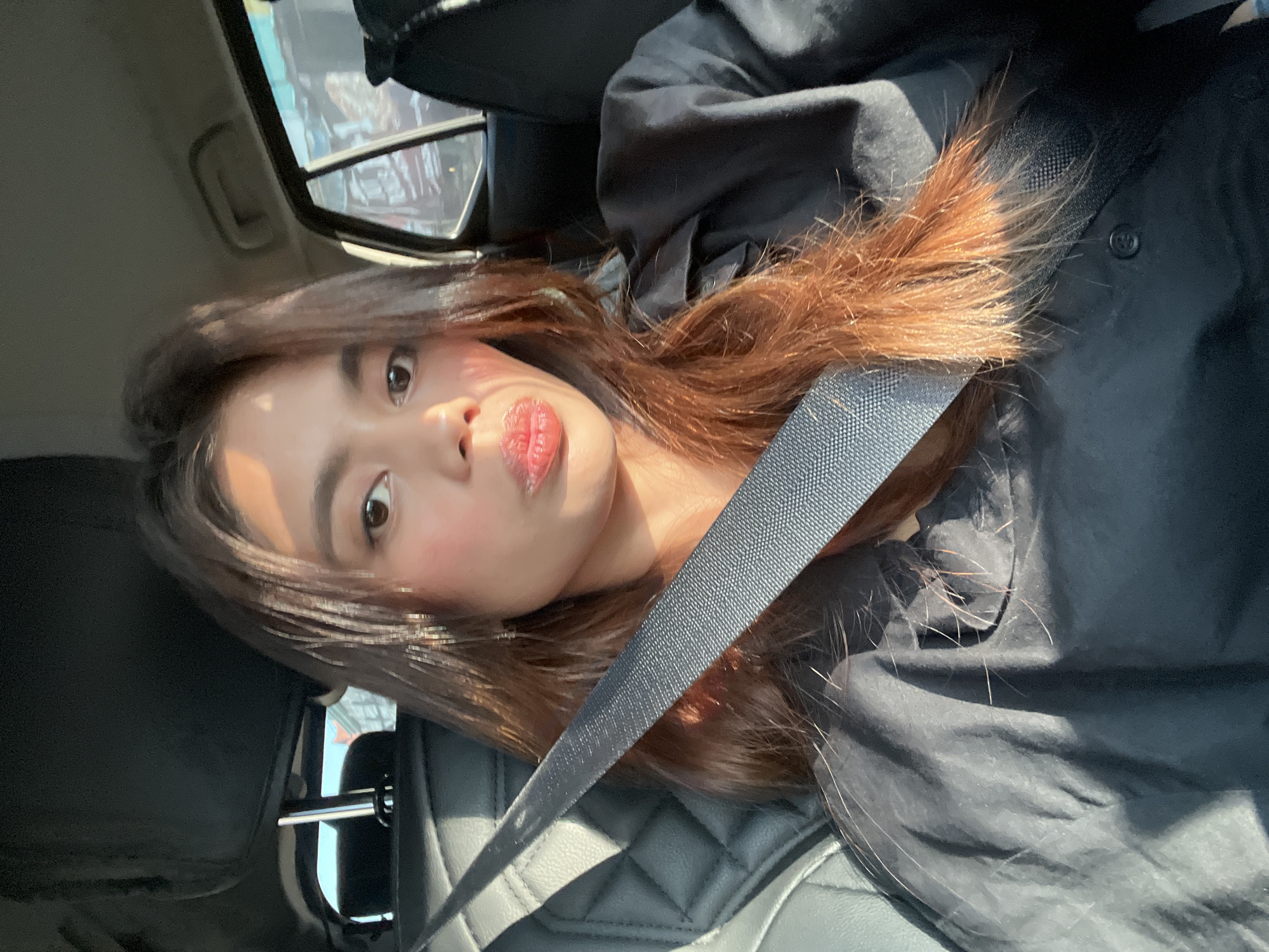 A solo female subject is depicted inside a passenger vehicle, located in Davao City, Philippines. She has long, dark brown hair with reddish undertones highlighted by direct sunlight. Her skin tone is light to medium, and she wears light facial makeup. Her lips are puckered, and she looks directly into the camera. She is secured by a black fabric seatbelt worn diagonally across her chest and is dressed in a dark, possibly black, long-sleeved collared shirt. The vehicle's interior features dark upholstery, with diamond-pattern stitching visible on the seat to her left. Strong sunlight enters from the right, casting defined shadows on her face and clothing. The exterior environment, visible through the rear window, is blurred, indicating an outdoor, likely urban or suburban, setting.