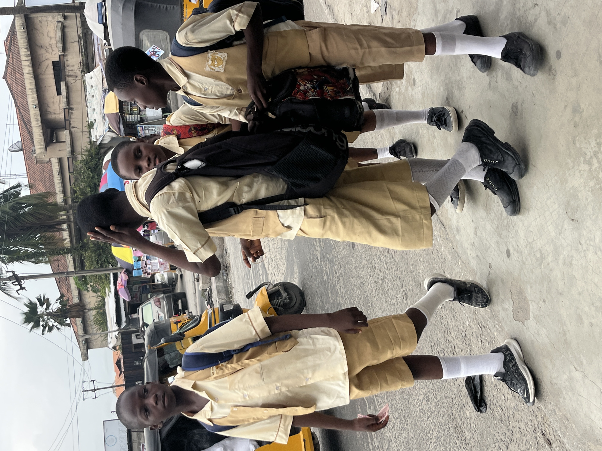 Four schoolboys in tan uniforms and white socks stand on a concrete sidewalk. The boys are of African descent. Three of them wear backpacks. The environment includes parked auto rickshaws, parked cars, buildings, power lines, trees, and street vendors. The location is likely Ado, Nigeria.