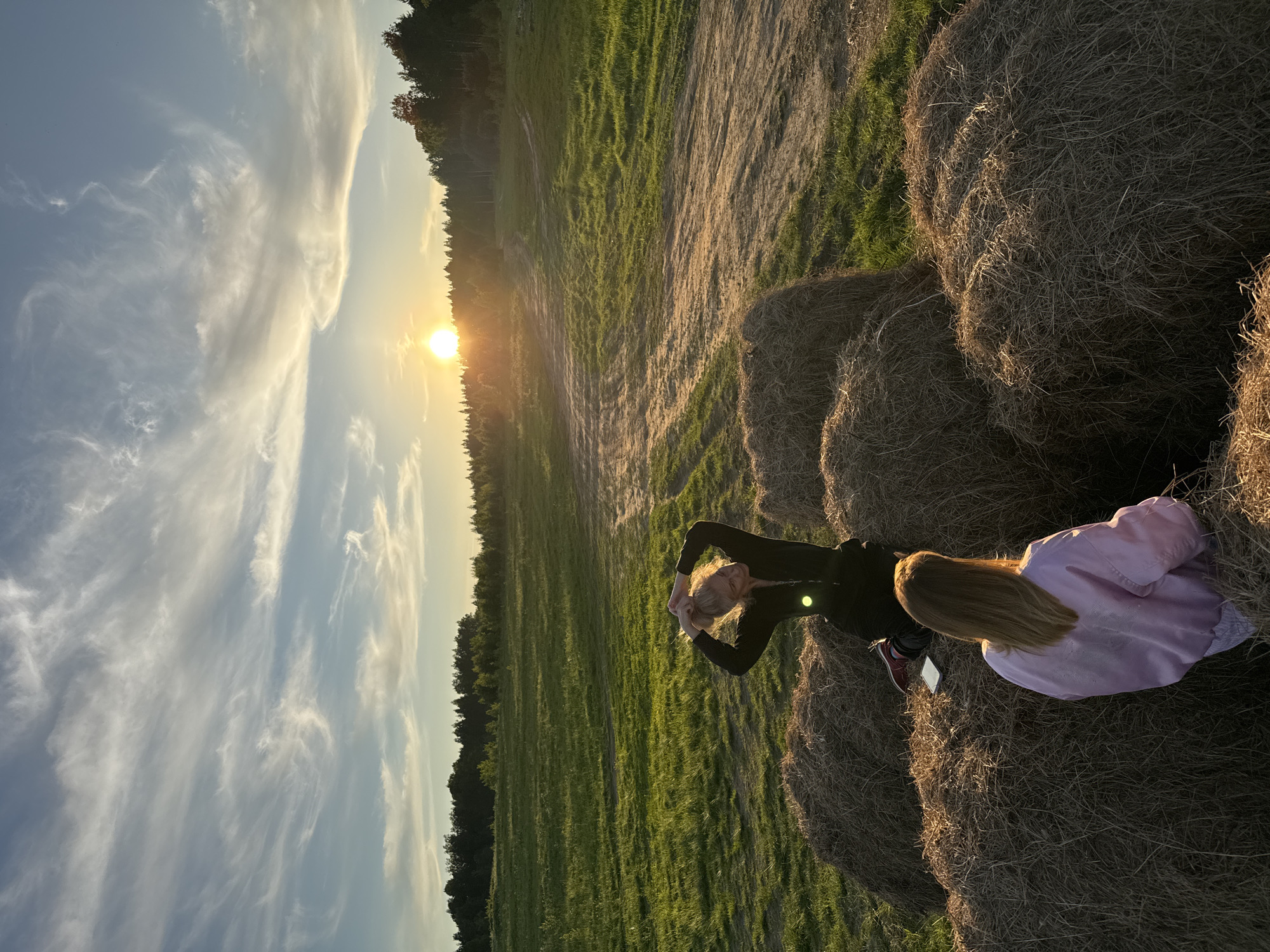 The image captures a serene outdoor scene at sunset in what appears to be a rural or agricultural setting, possibly in Стеклянный, Russia. The warm, golden light of the setting sun bathes the landscape, creating long shadows and highlighting textures.
In the foreground and midground, two individuals are seated on large, round hay bales. One person, positioned further from the camera, has long blonde hair and is wearing a dark long-sleeved top. They have their arms raised and clasped behind their head, looking towards the setting sun, possibly stretching or enjoying the view. The second person, closer to the camera and seen from behind, also has long blonde hair and is wearing a light purple jacket. A white smartphone or similar device rests on the hay bale next to them.
The hay bales are prominently featured, providing a rustic seating area. Beyond the bales, a vast, green grassy field stretches into the distance, traversed by light dirt paths. A dense line of trees forms the horizon, silhouetted against the bright sky where the sun is low.
The sky is a pale blue, adorned with wispy, elongated cirrus clouds that catch the golden light from the sun, creating a dynamic and picturesque backdrop. The overall mood is peaceful and contemplative, suggesting a moment of relaxation at the end of the day. There is no visible text in the image.