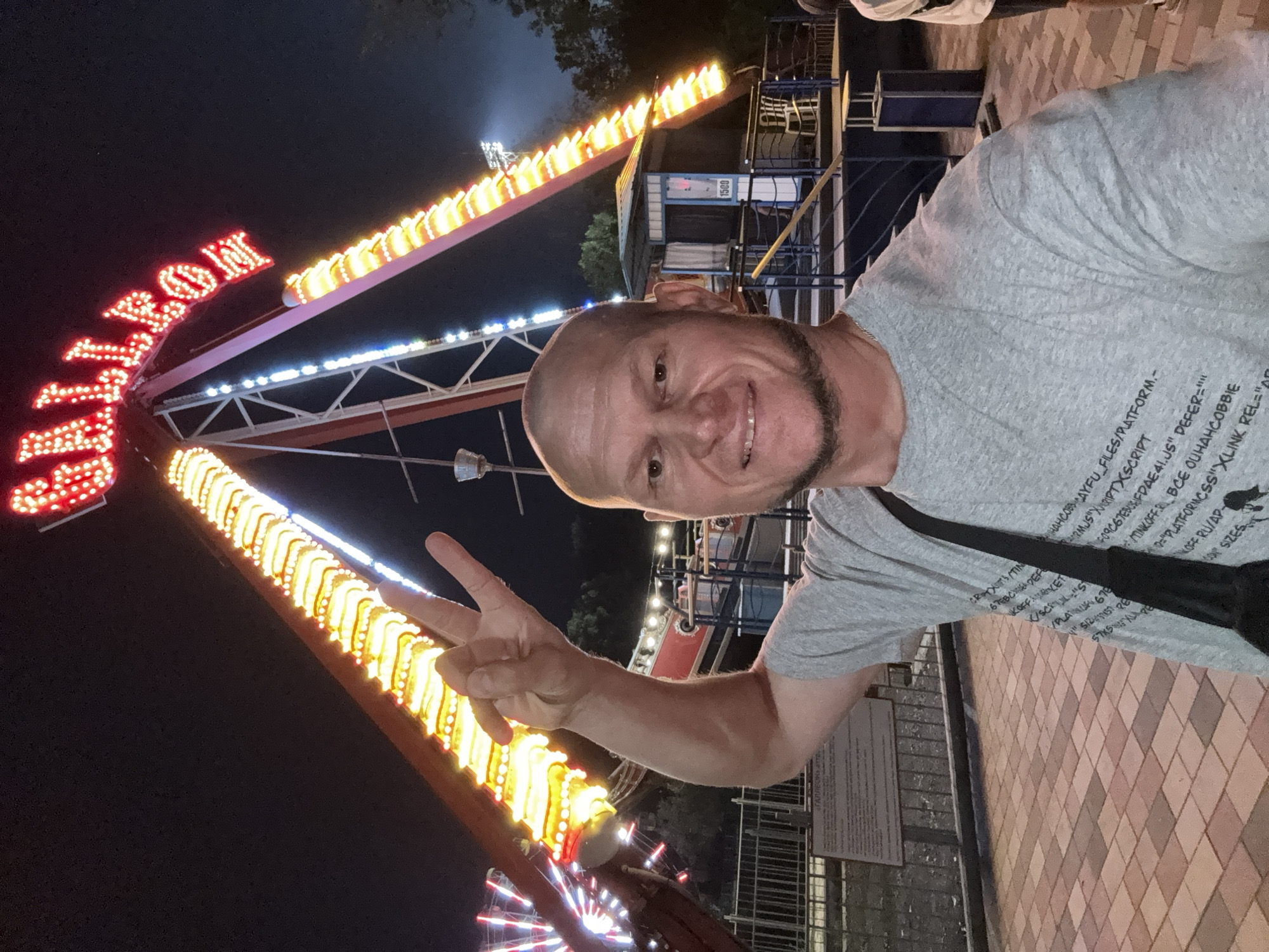 A bald, Caucasian man with a goatee and a grey t-shirt is taking a selfie at night in an amusement park in Oskemen, Kazakhstan. He is making a peace sign with his left hand. Behind him, a large amusement park ride, a pendulum ride shaped like a pirate ship, is illuminated with bright yellow and red lights. The ride's name, "GALLEON," is spelled out in red neon lights at the top. The man is standing on a paved walkway with cobblestone patterns. In the background, there are more amusement park attractions, including a Ferris wheel, and a control booth for the ride. The atmosphere is festive and colorful, typical of a nighttime amusement park setting.