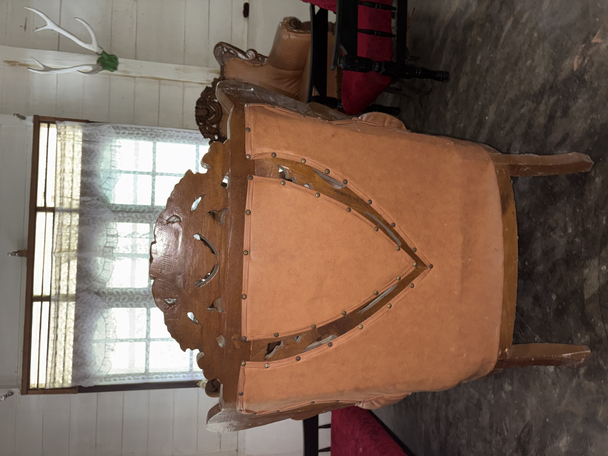The image shows an interior scene of a room, likely a living area, with antique furniture. The focal point is the back of a large, ornate wooden armchair with tan upholstery. The upholstery is studded with small metal tacks, forming a decorative pattern. Behind this armchair, other furniture pieces are visible, including a dark-colored chair with a red seat and another brown upholstered chair. A window with lace curtains is present in the background. On the wall to the right, a set of white deer antlers is mounted. The flooring appears to be concrete. The overall impression is that of a room decorated with vintage and possibly traditional Indonesian furnishings. There is no visible indication of the city of Beunyot, Indonesia in the image itself.