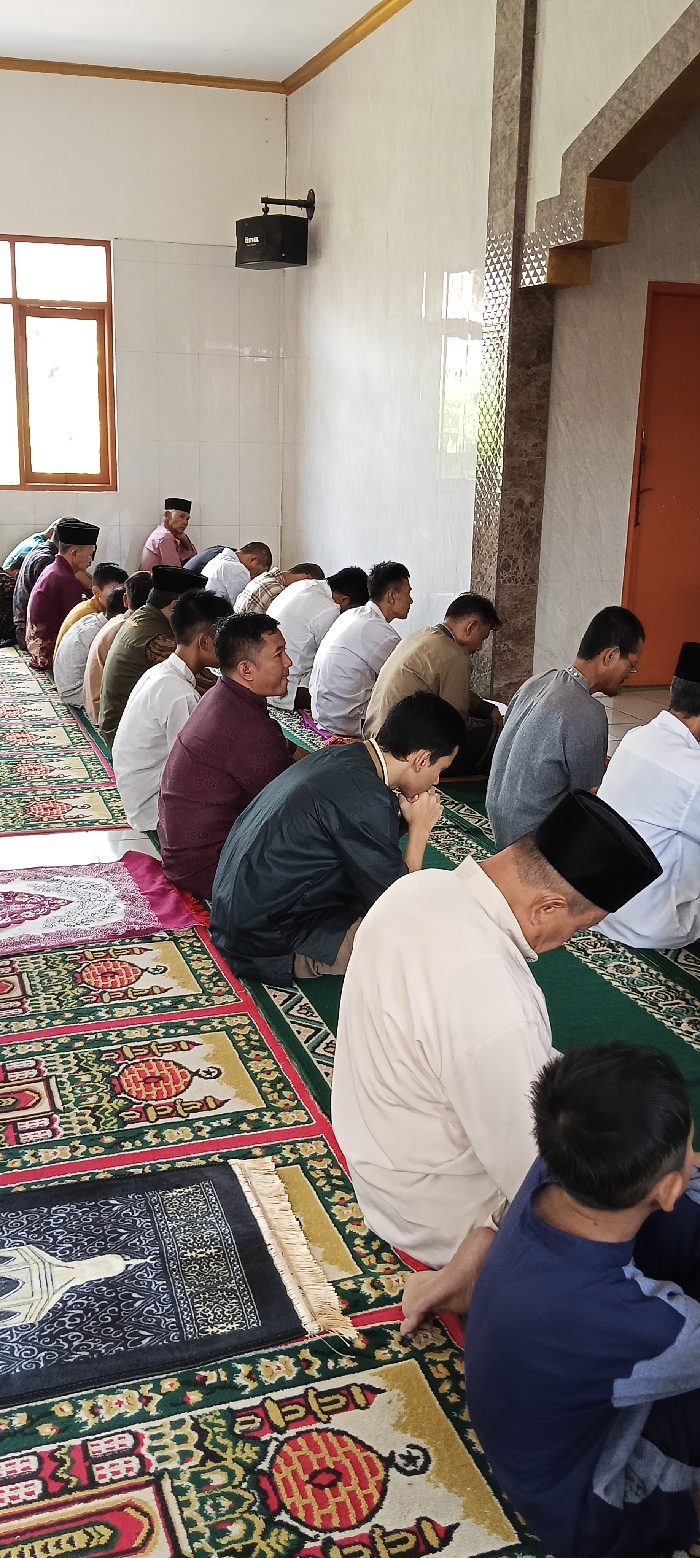 The image shows a group of men in a mosque located in Kecamatan Kutawaringin, Indonesia. They are sitting on prayer rugs with their heads bowed in prayer. The men are wearing traditional clothing and some have black skullcaps on their heads. The room is clean and well-lit, with white walls and a tiled floor. There is a window on the left side of the room that shows trees and a building in the distance. A speaker is mounted on the wall in the top left corner of the room. There is a door on the right side of the room that leads to another room. The men are engaged in silent prayer, their heads bowed in unison, and the atmosphere is calm and serene. There is a young boy sitting on the floor, with his back to the camera, watching the men praying. The mosque is decorated with traditional Islamic patterns and designs.