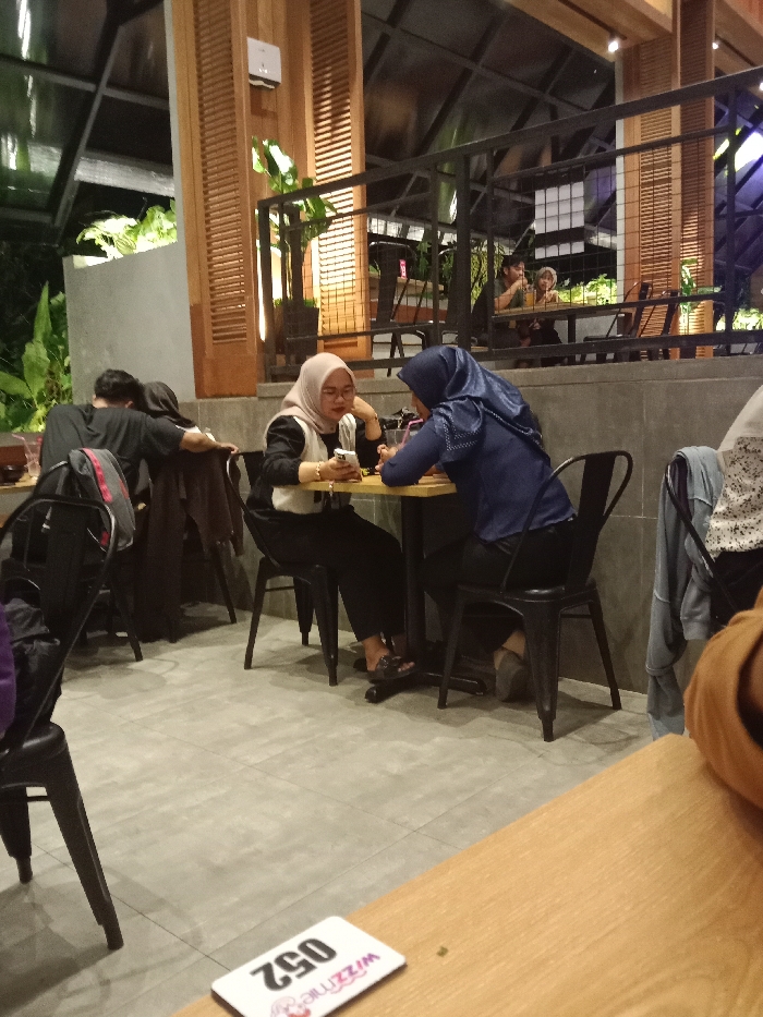 The image shows a scene inside a restaurant in Kecamatan Ngaliyan, Indonesia. There are four people in the image. Two women are sitting at a table near the center of the frame, talking to each other. One of the women is wearing a white hijab and the other is wearing a blue hijab. The woman in the white hijab is looking down at her phone, while the woman in the blue hijab is looking at her. A man wearing a black shirt is seated in the left background, he appears to be asleep, and another person is not fully visible. The table is made of wood and there are four black chairs around it. There are a few other tables and chairs in the background, which are empty. The restaurant is modern and the walls are covered in tiles. The floor is made of concrete. There is a wooden slatted wall in the background, and the glass ceiling is open.