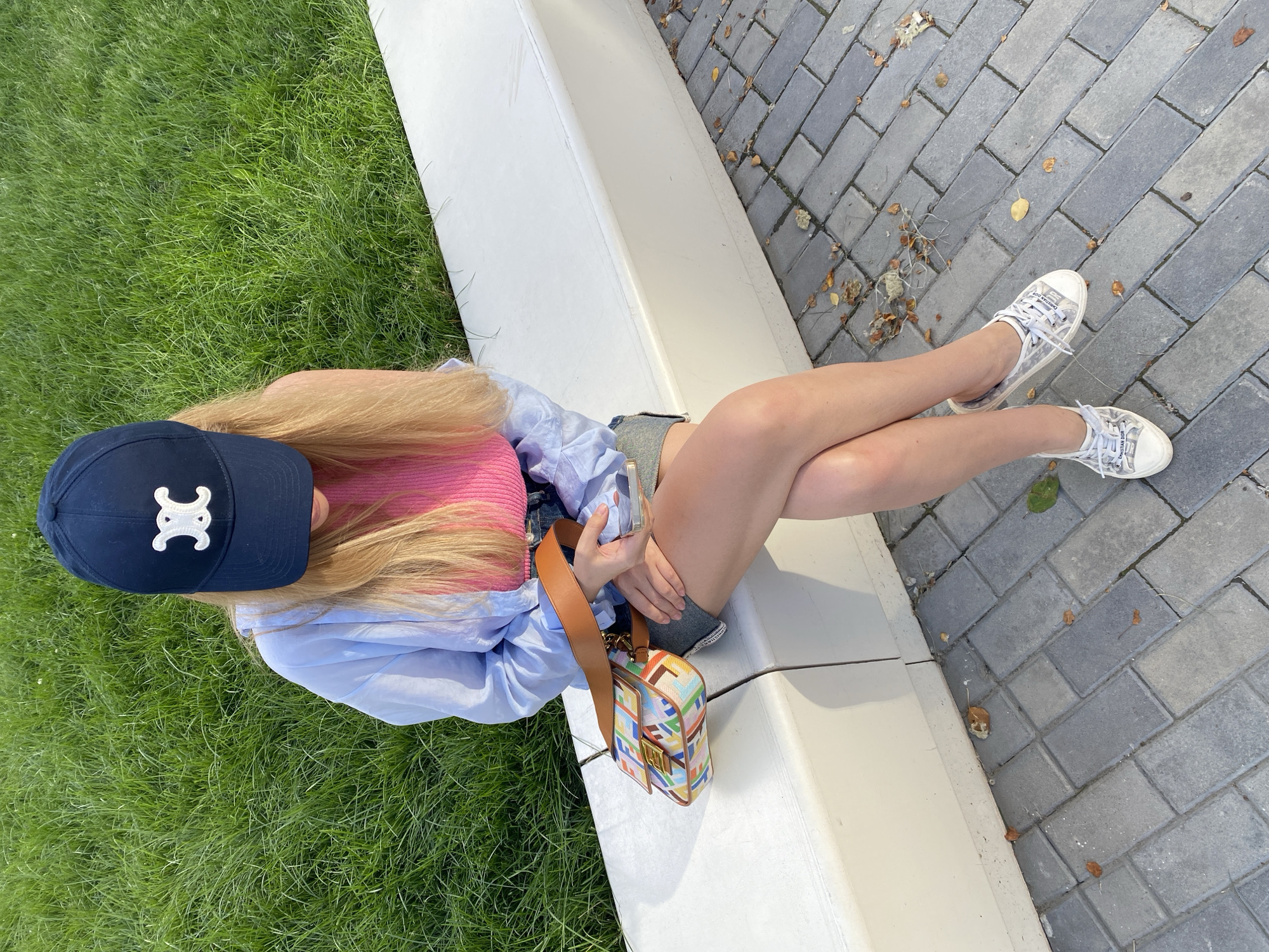 A young Caucasian woman with blonde hair, wearing a navy blue baseball cap with a white logo, a pink ribbed tank top, an open light blue button-down shirt, and denim shorts, is sitting on a white concrete bench. She is holding a smartphone and looking down at it. A colorful, patterned handbag with a brown leather strap is resting on the bench next to her. She is wearing white low-top sneakers with visible branding. The background consists of a grassy area and a paved walkway made of grey rectangular stones. Fallen leaves are scattered on the ground. The scene appears to be an outdoor public space in Yekaterinburg, Russia.