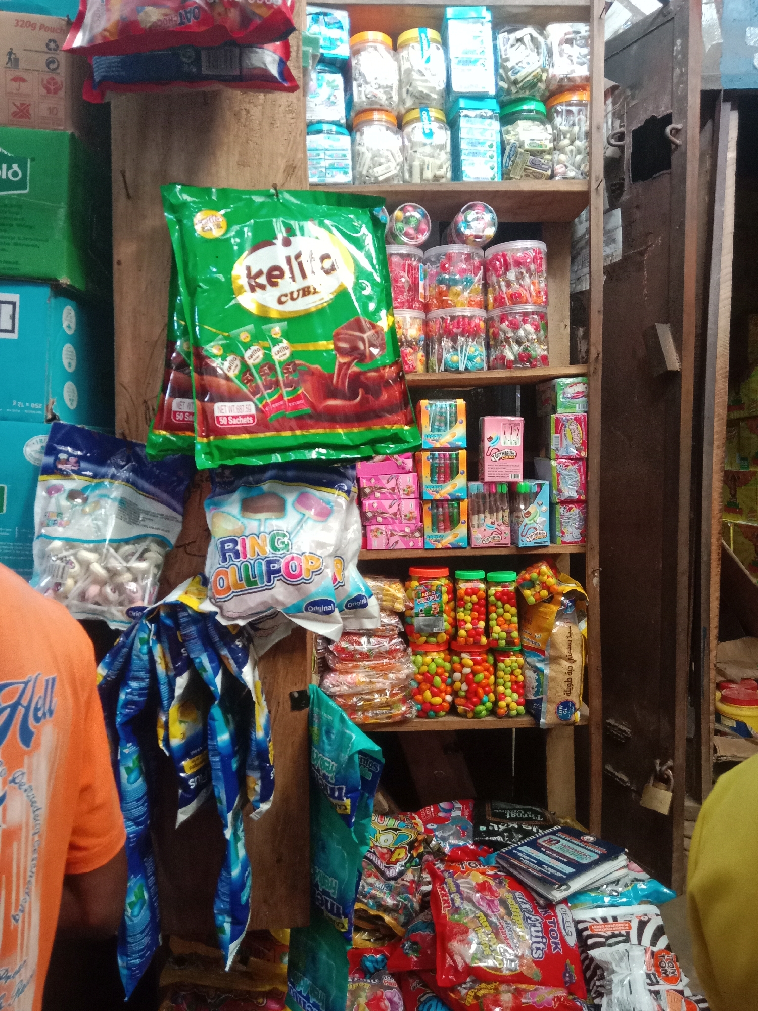 The image shows a retail display of candy and sweets in Onitsha, Nigeria. The merchandise is arranged on a wooden shelf and on the floor. The shelf contains jars of candy, boxes of sweets, and bags of lollipops and hard candies. Floor displays include bags of lollipops, hard candies, and several notebooks. The scene is located in a store where the display is situated between two vertical boards. A person wearing an orange shirt is seen on the left, while another person wearing a yellow shirt is seen on the right. A padlock secures something in the background.