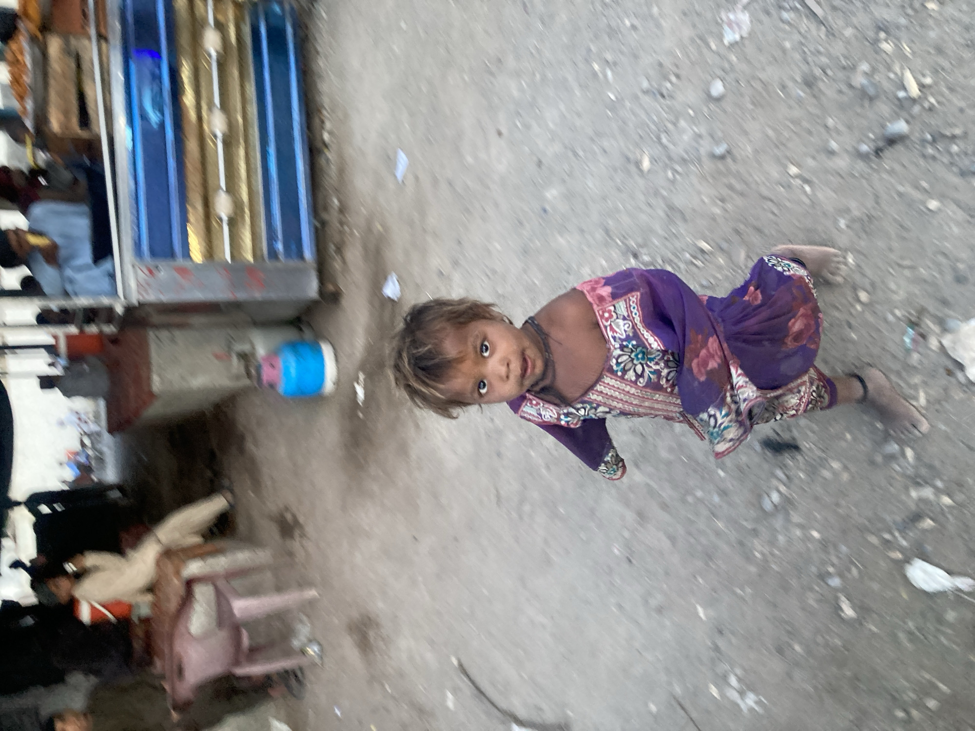 This candid, angled photograph captures a young child looking upwards at the camera in an outdoor, unpaved setting in Uthal, Pakistan. The child, appearing to be a toddler or very young, is positioned in the lower portion of the frame, their head slightly tilted. They have light brown hair and wide, dark eyes, wearing a vibrant purple dress adorned with intricate floral and geometric patterns in pink, white, and blue hues. A simple, dark necklace is visible around their neck, and their bare feet are visible on the ground.
The immediate foreground and much of the background consist of a dusty, grey, unpaved surface, likely a dirt or gravel path, strewn with small stones and various pieces of light-colored debris. To the left of the child, a section of a structure or stall is visible, characterized by vertical blue and golden panels. Further into the background on the left, other people are faintly visible, one person appears to be seated or resting on a light-colored chair. A blue container or bottle is also on the ground near these background elements. The lighting suggests it is daytime, possibly overcast or late afternoon, as there are no harsh shadows. There is no visible text. The scene conveys a sense of everyday life in a modest, possibly informal, street or alley setting.