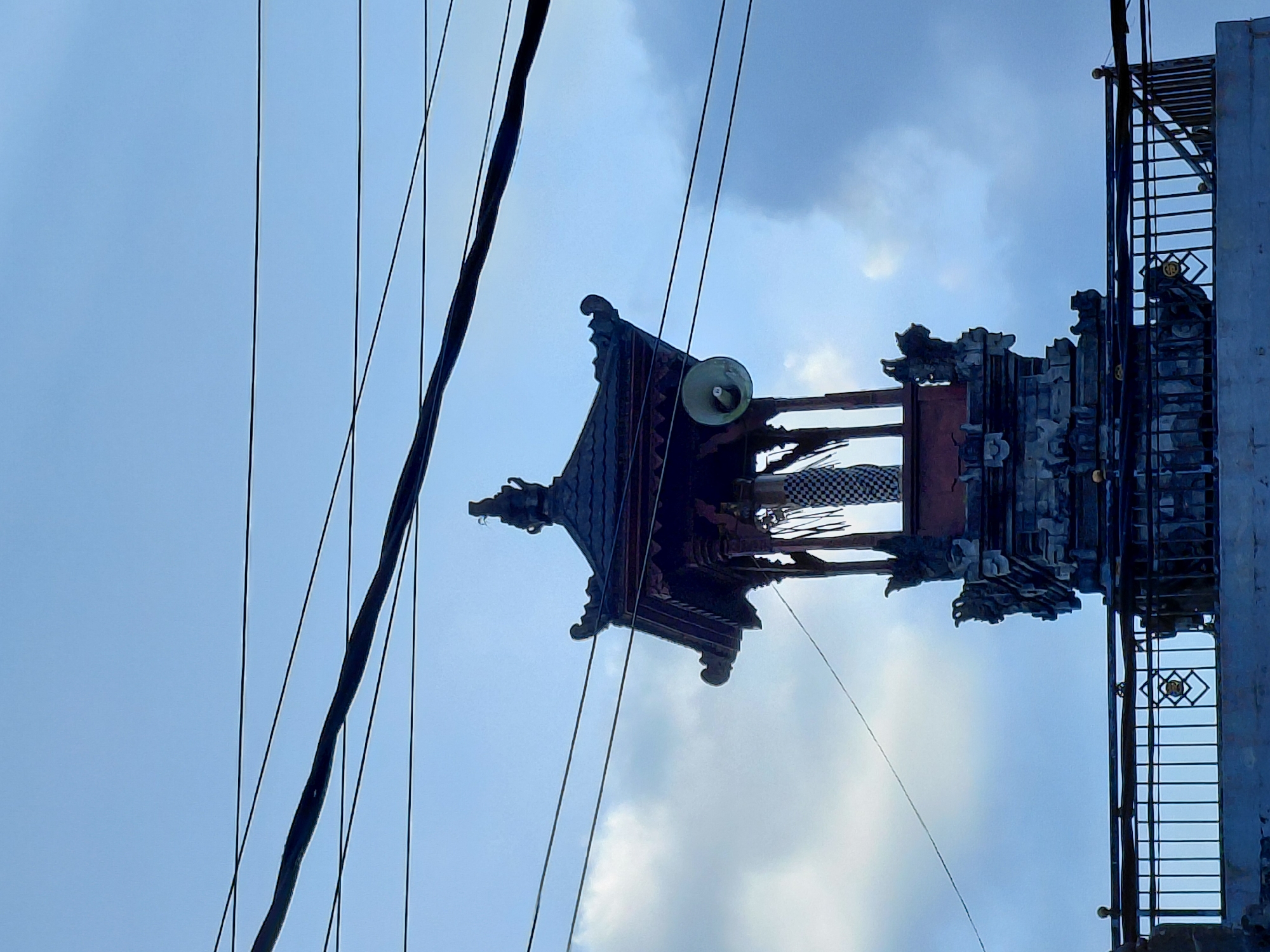 A traditional Balinese temple structure stands against a cloudy blue sky, with several black power lines crossing the frame. The temple features multiple tiers, with intricate stone carvings and a dark, multi-layered roof. A white speaker is mounted on the side of the upper section of the temple. A black metal railing with decorative elements runs along the base of the structure. The image appears to be taken in the city of Sukasada, Indonesia, given the architectural style and context. There are no people visible in the image.