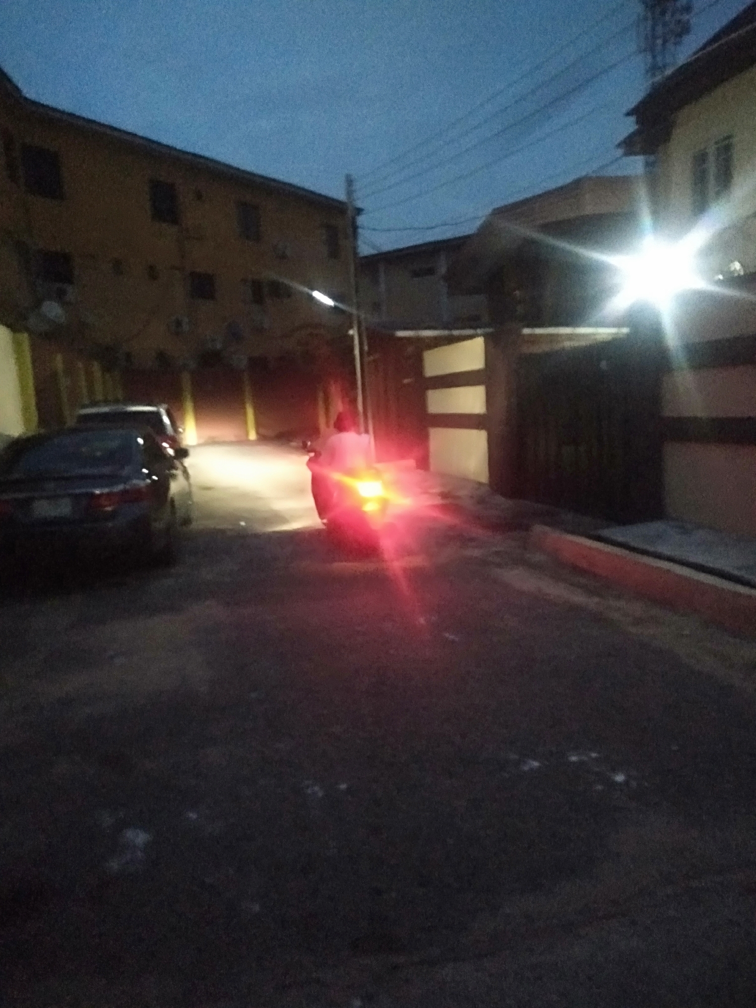 A street in Unknown, Unknown is dimly lit by a street lamp and the tail lights of a motorcycle. Three cars are parked on the left side of the street. A person is riding the motorcycle, moving away from the viewer. Buildings line the street on both sides. The time of day is likely dusk or night.