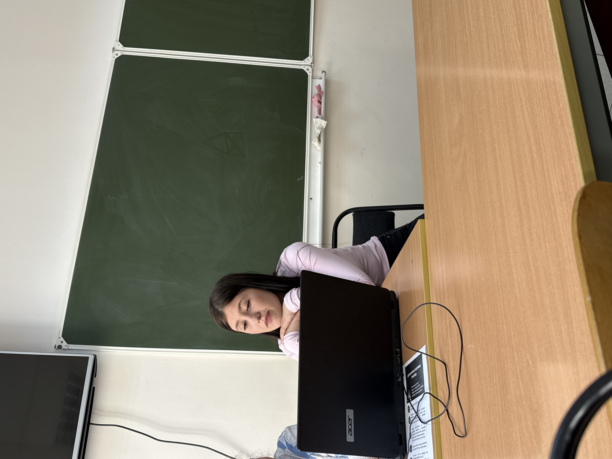 A medium shot shows a young woman seated at a desk in what appears to be a classroom in Chaikovsky, Russia. She is leaning on her arms and looking toward a black Acer laptop in front of her. She has dark hair, light skin, and is wearing a light pink long-sleeved shirt.
Behind her is a green chalkboard with a metal frame, mounted on a white wall. To the left, a flat-screen monitor is visible. To the right, a light brown wooden desk extends into the frame. The woman is seated on a black chair. A cable for the laptop and some papers are also on the desk.