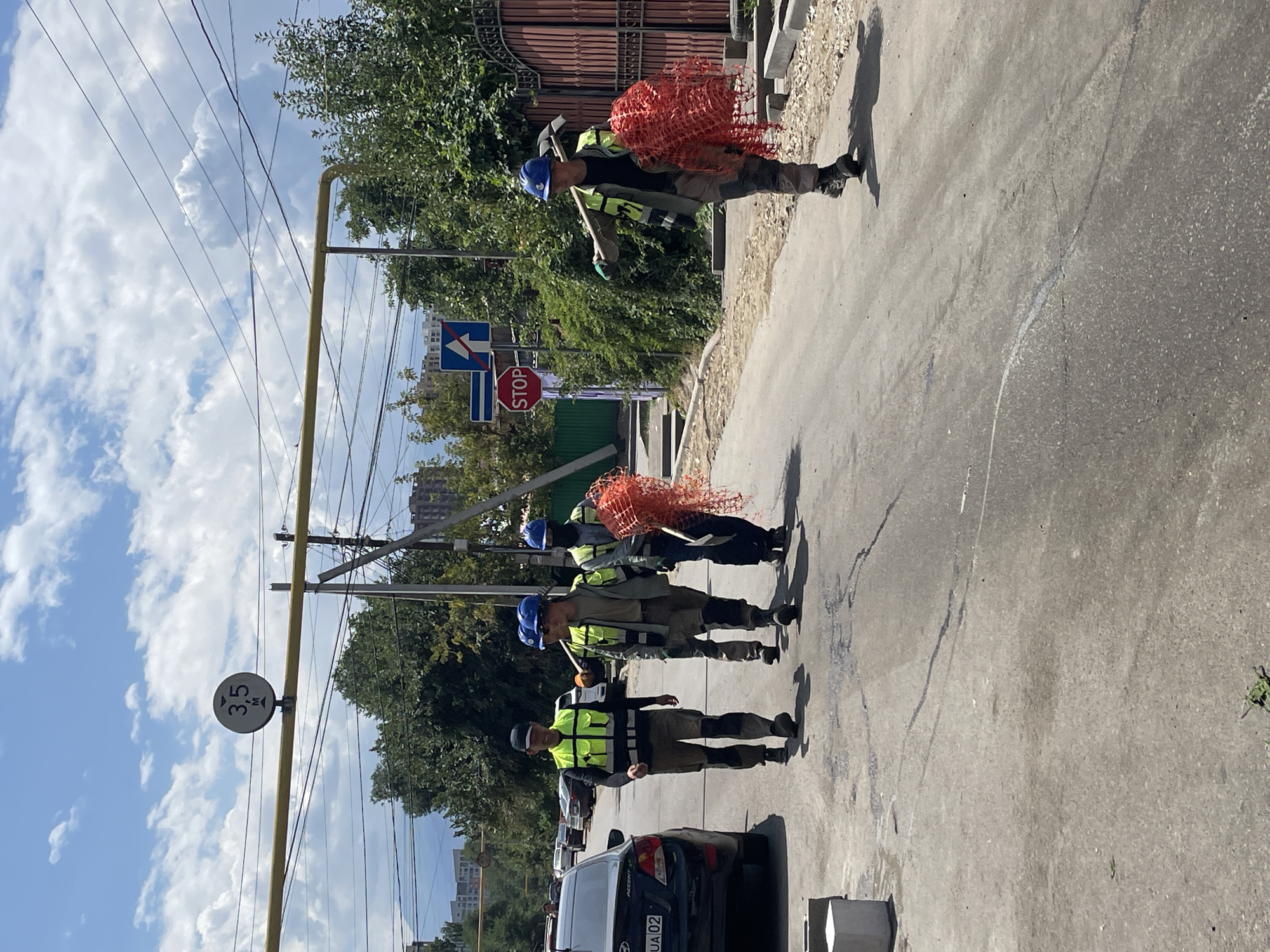Four individuals wearing high-visibility vests and hard hats are walking on a paved road. Two of the individuals are carrying shovels, and one is carrying a bundle of orange netting. The individuals are walking in the same direction. In the background, there are cars, buildings, trees, and power lines. A speed limit sign showing "3,5 M" is visible above the road, and a "STOP" sign is posted on the right side of the road. The scene appears to be in an urban or suburban area, possibly Almaty, Kazakhstan, given the context provided. The road is undergoing some form of construction or repair, with debris visible on the side. The sky is partly cloudy.