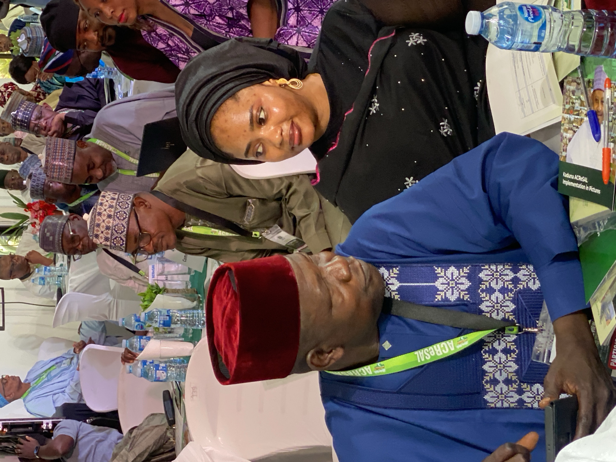 The image shows a conference setting in what appears to be Kwarau, Nigeria. Several people are seated at tables, some wearing traditional attire. A woman in a black headscarf and dress with floral accents is seated at the foreground of the table. A man in a blue outfit and a red hat is sleeping, with an accreditation lanyard around his neck labeled "ACReSAL". A book titled "Kaduna ACReSAL Implementation in Pictures" is visible on the table next to a bottle of water.