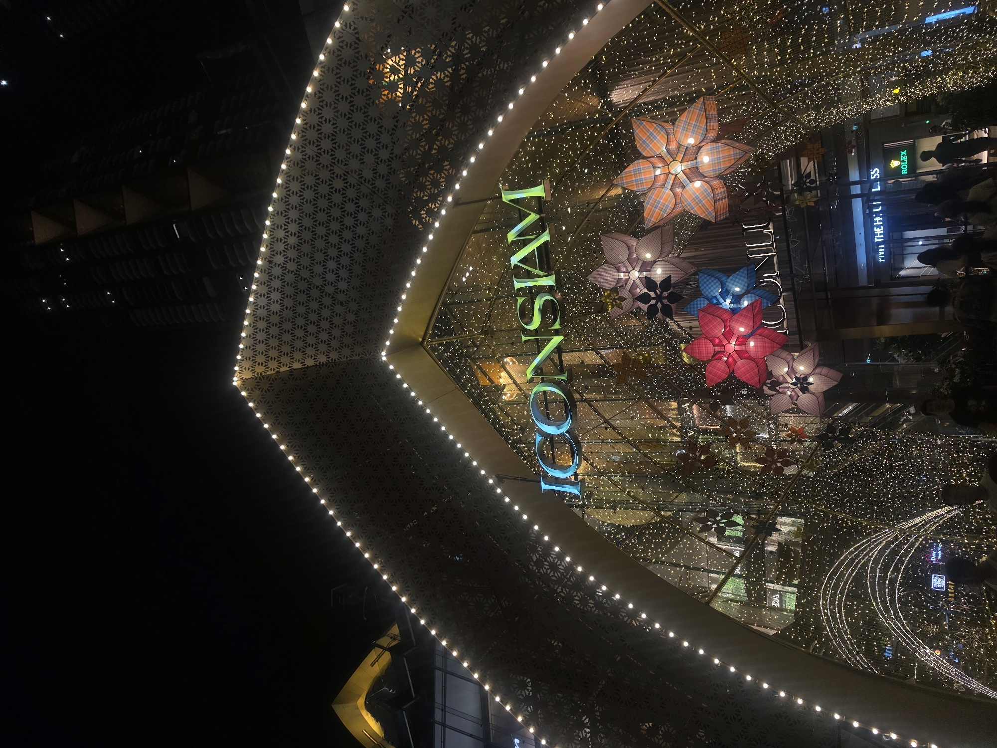 The image captures a nighttime view of the ICONSIAM shopping mall in Bangkok, Thailand, known for its riverside location and luxury retail offerings. The mall's entrance is decorated with numerous twinkling lights that create a festive atmosphere. The large "ICONSIAM" sign, illuminated in green and blue, is prominently displayed. Oversized, decorative flower-shaped lanterns in various colors hang from the ceiling. People are visible inside the mall, suggesting activity and commerce. The architecture incorporates geometric patterns, and the area is well-lit, highlighting the mall's modern design.