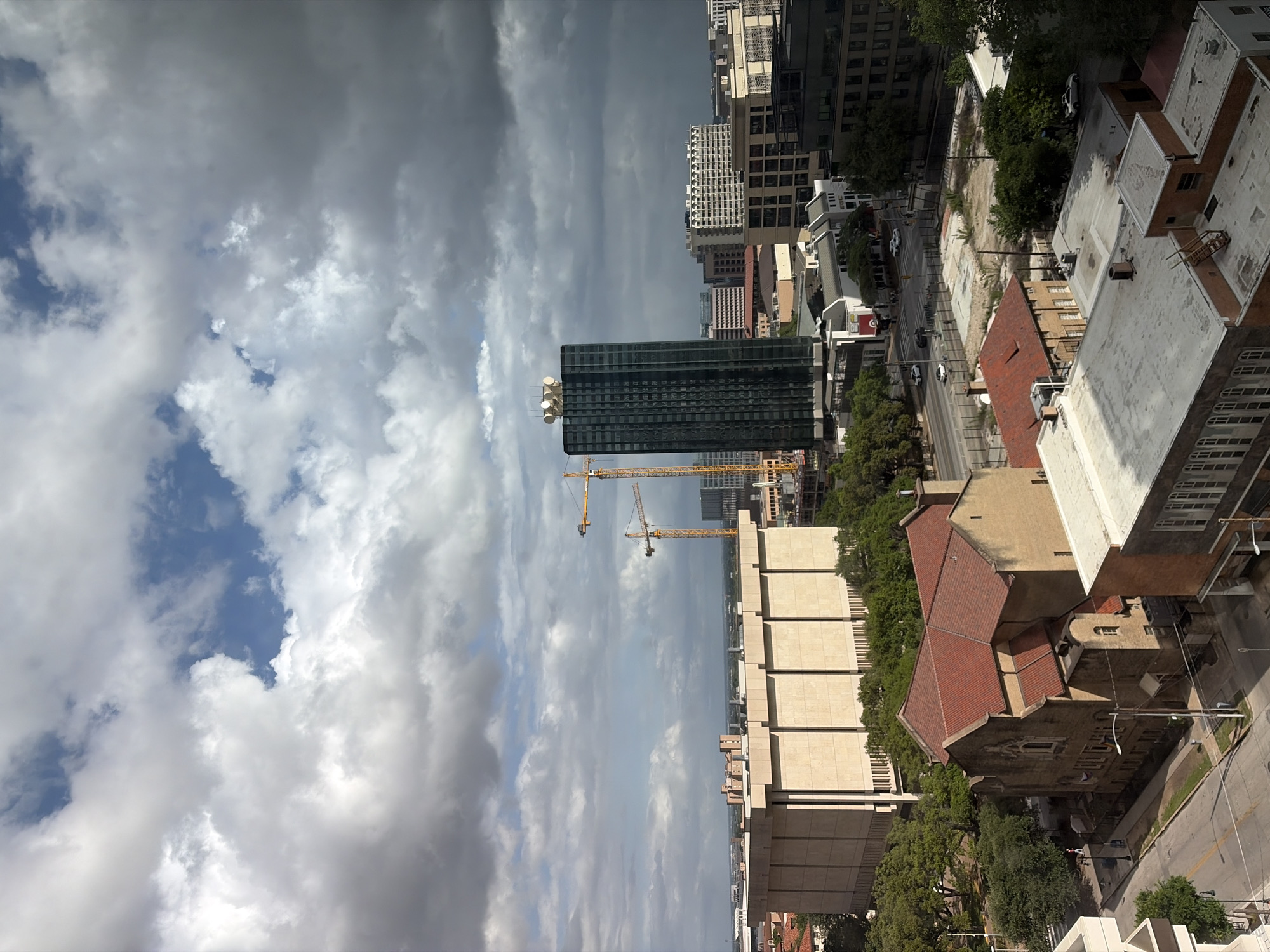 The image shows a cityscape view in Austin, Texas, United States of America, under a sky with dramatic cloud formations. A tall, dark glass skyscraper dominates the middle ground, partially obscured by two yellow construction cranes. Adjacent to the skyscraper is a light-colored, multi-story building with a modular design. Closer to the right, there is a building with a red-tiled roof and adjacent buildings of beige brick and cream color with flat roofs. Beyond the skyscraper, other high-rise buildings are visible in the distance. A street runs between the buildings in the foreground, with some vehicles visible. The overall scene suggests a mix of construction, urban landscape, and variable weather conditions.