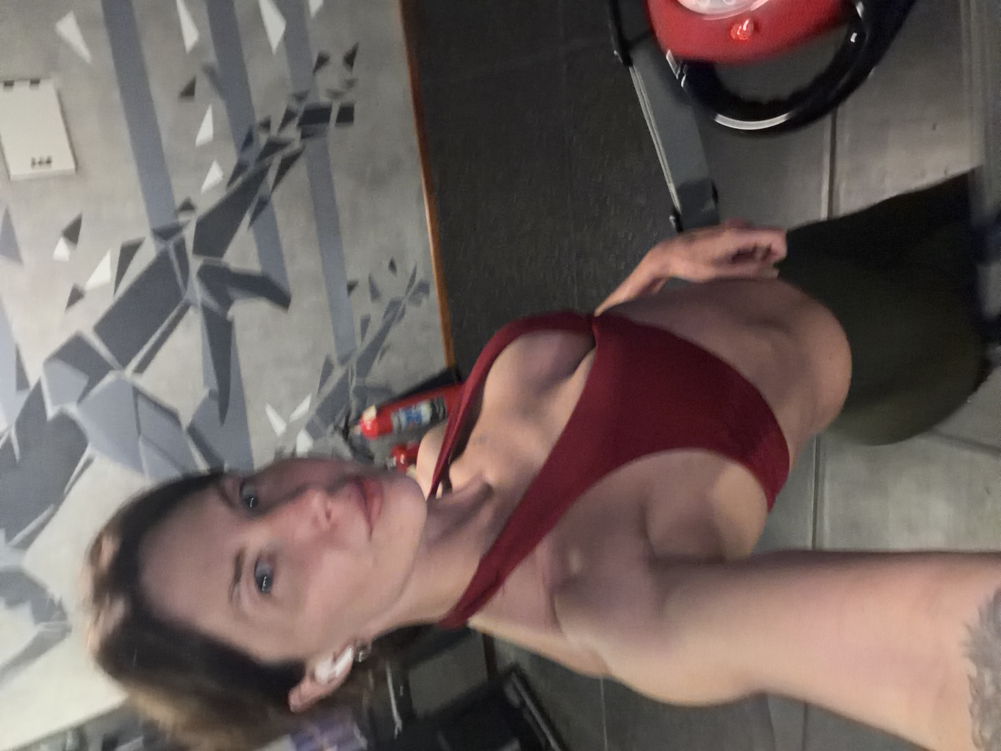 In the city of Dong Ninh Hoa Ward, Vietnam, a woman is captured in a selfie at a gym. She is wearing a dark red halter-neck sports bra and dark green leggings. Her short brown hair is neatly styled, and she has white earbuds in her ears. The background reveals gym equipment, including a treadmill, and a wall with a geometric abstract mural. The overall environment appears to be a fitness facility, possibly a personal training studio or a commercial gym. The lighting is dim, typical of indoor gym settings.