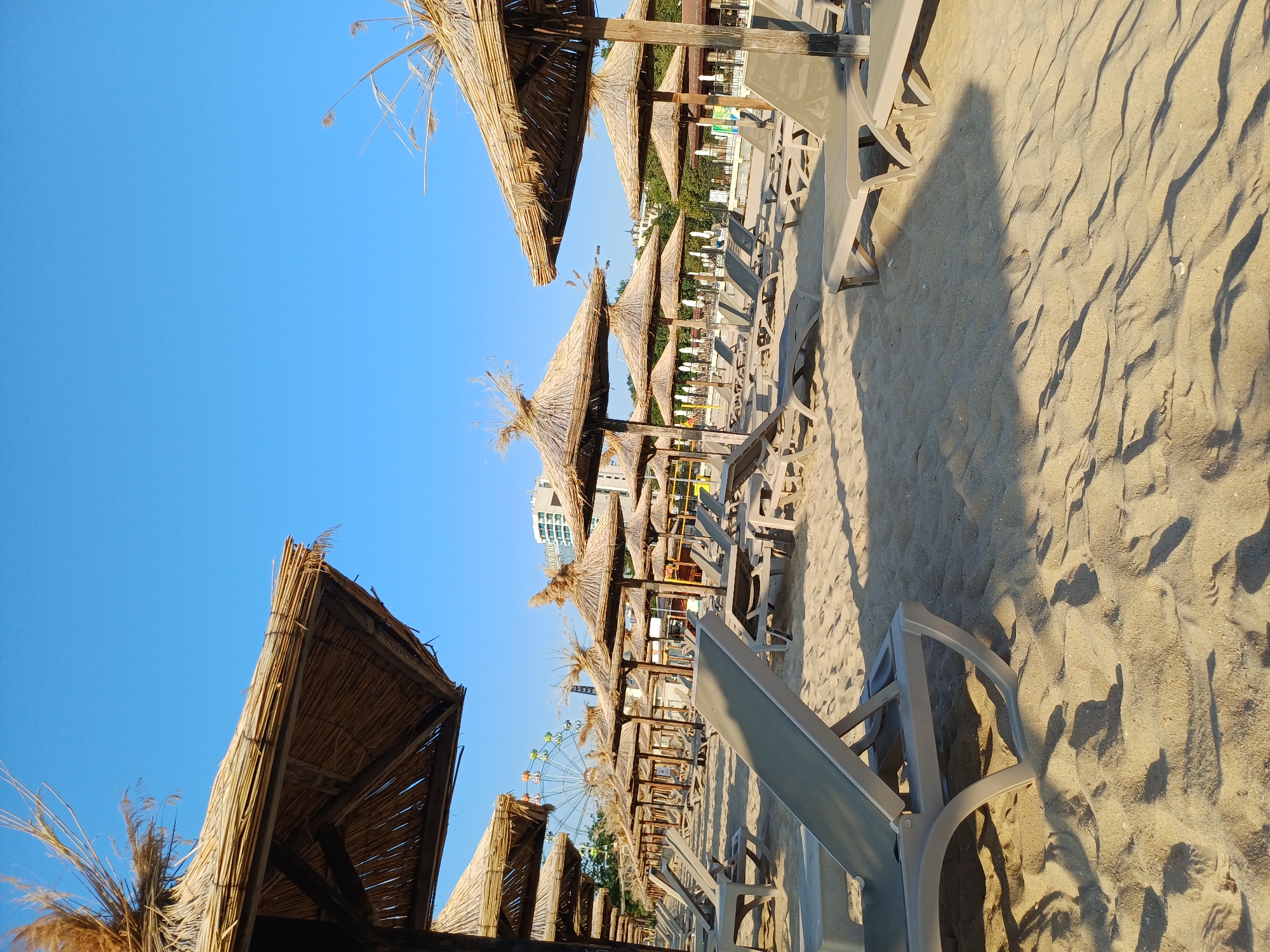 The image depicts a beach scene in Varna, Bulgaria, characterized by rows of empty lounge chairs and thatched umbrellas under a clear blue sky. The sand is light in color and shows the texture of footprints and ripples. In the background, a Ferris wheel is visible, along with several buildings, suggesting an urban coastal setting. The lighting indicates it is likely daytime, with shadows cast by the umbrellas and chairs. The overall atmosphere is one of a quiet, prepared beach, possibly before or after peak hours.