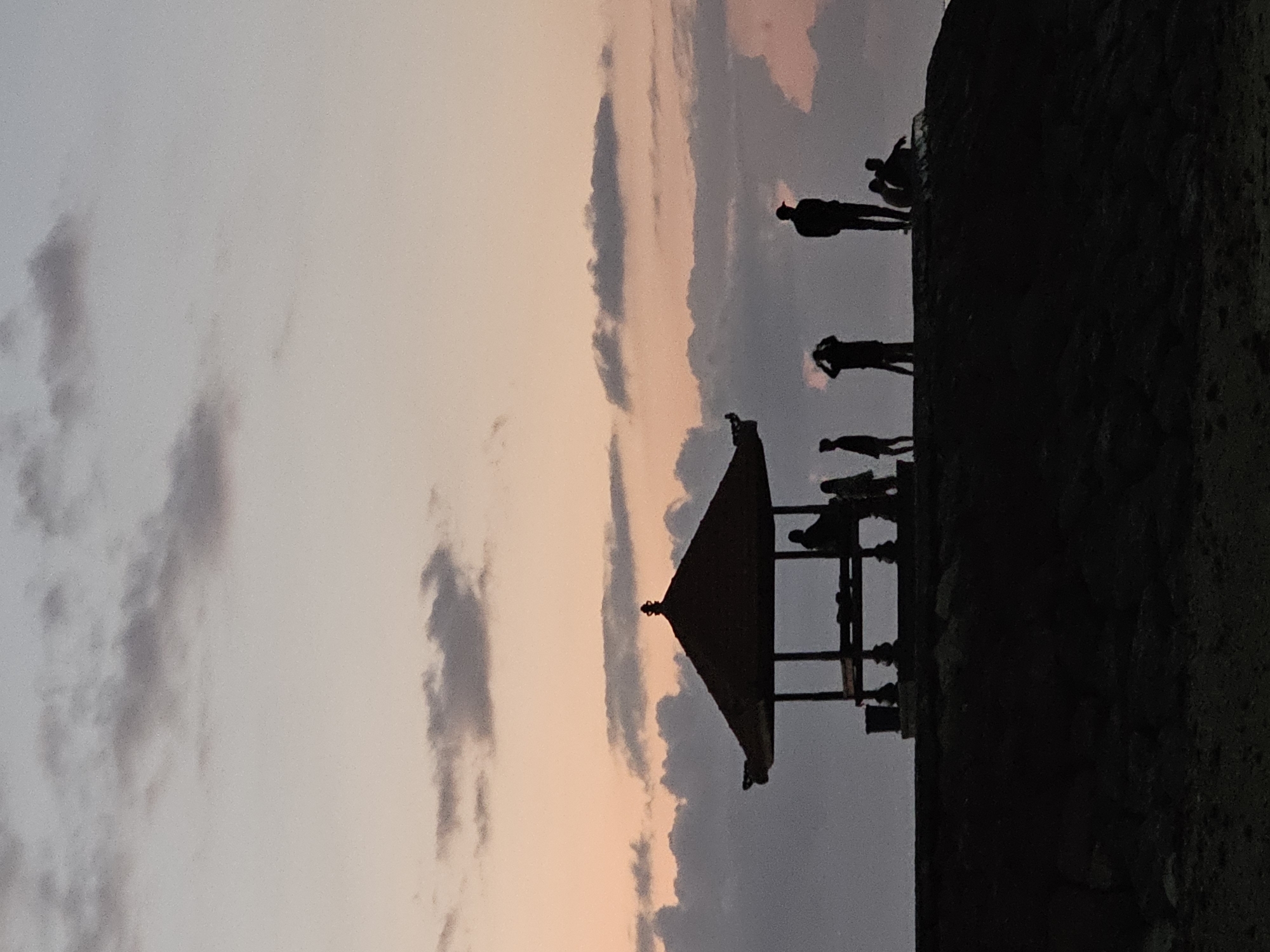 A silhouetted group of people are gathered near a gazebo on a hilltop overlooking the ocean at sunset in Denpasar, Indonesia. The sky is filled with hues of pink, orange, and purple, with scattered clouds. The gazebo, with its distinctive pointed roof, is located on the left side of the image. Several individuals are visible in and around the gazebo, some appearing to be sitting or standing. To the right of the gazebo, more people are silhouetted against the sky, some standing and some sitting. The foreground is dark and appears to be rocky terrain. The overall scene depicts a tranquil evening gathering at a scenic location.