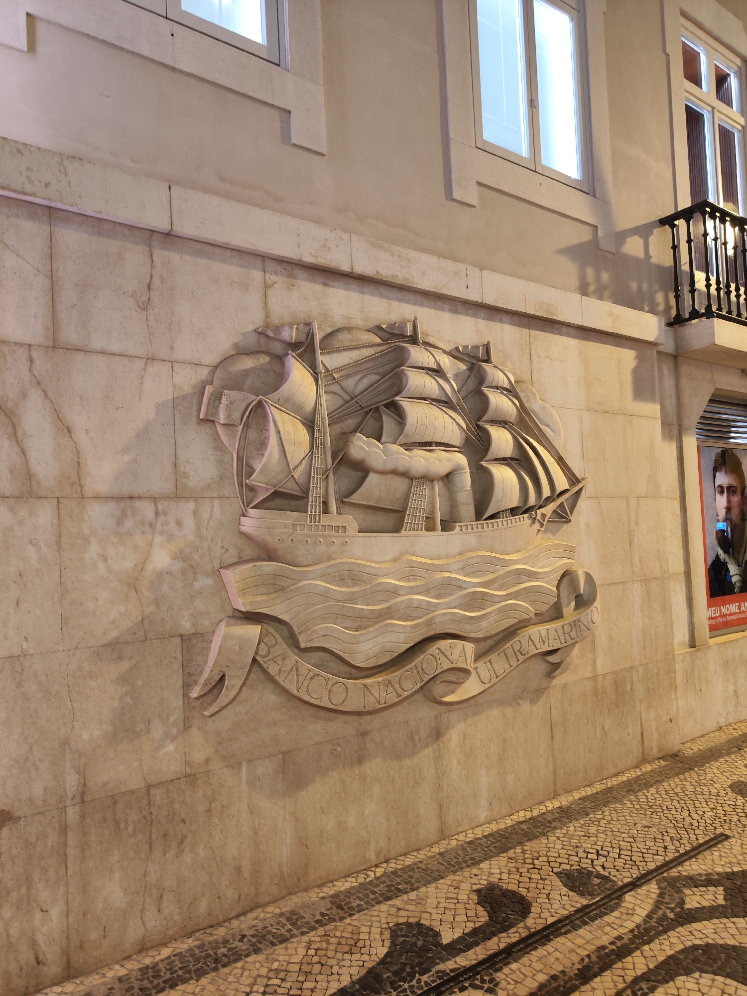 The image displays an evening street scene in Lisbon, Portugal, featuring a prominent stone bas-relief on the facade of a building. The central element is an elaborate carving of a large, three-masted sailing ship with multiple sails unfurled, depicted atop stylized waves. A banner beneath the ship carries the inscription "BANCO NACIONAL ULTRAMARINO" in capital letters.
The carving is affixed to a light-colored, possibly marble or limestone, building wall, which exhibits natural veining and some signs of age. Above the carving, the building features several rectangular windows, glowing brightly from internal illumination, indicating that it is evening or night. To the right, a black wrought-iron balcony extends from an upper floor.
At street level, there is a traditional Portuguese pavement (calçada portuguesa) made of small, irregularly shaped light and dark-colored stone tiles, arranged to form decorative patterns, including swirling shapes. Artificial light sources cast long, dark shadows of unseen street furniture or architecture across this patterned sidewalk.
On the far right, a poster or advertisement is visible inside a display window. It features a close-up portrait of a man, and partial text can be deciphered, reading "MEU NOME A..." (likely "MEU NOME É...") and "fotografia de Teresa Cab...". The scene evokes a blend of historical architecture and urban ambiance under artificial lighting.