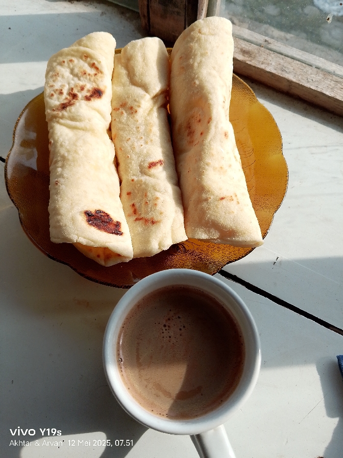 Three flatbreads are arranged on a brown, scalloped plate. The plate is on a white surface next to a white cup of coffee. The flatbreads are slightly browned from cooking. The coffee is dark brown and has a frothy top. The scene is in a home in Anjatan, Indonesia, at the coordinates -6.419 latitude, 107.978 longitude. There is a window in the background with a wood frame. The image was taken on May 12, 2023 at 07:51. The image was taken with a Vivo Y19s phone. There are no people in the image.