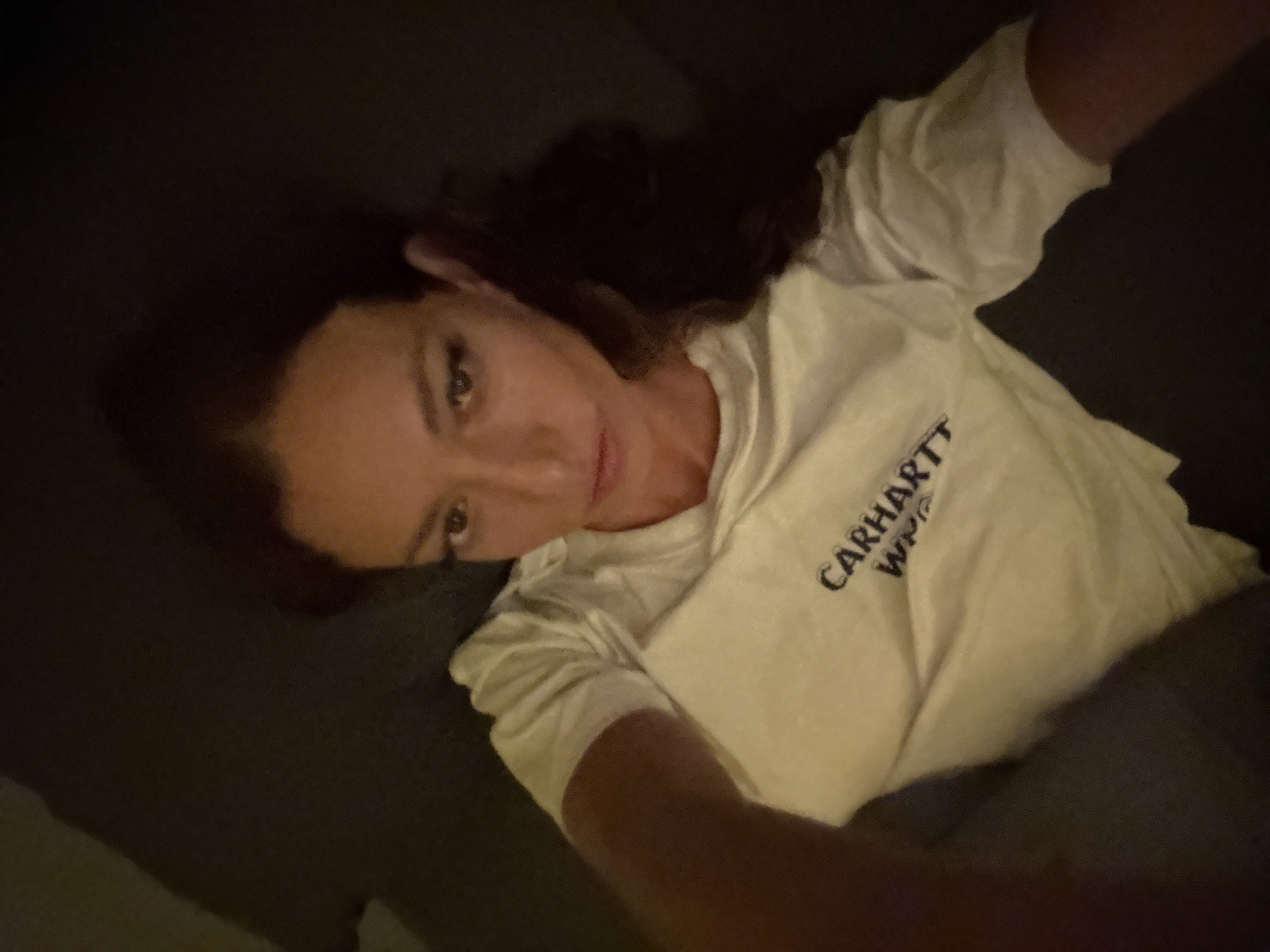 A woman with dark hair is lying down, wearing a white t-shirt with the word "CARHARTT" printed on it. The environment is dimly lit, and she appears to be on a dark gray surface, possibly a couch or bed. The location in Baan Chaweng, Thailand is not evident from the visual information provided. There are no actions or interactions visible, and the scene is static.