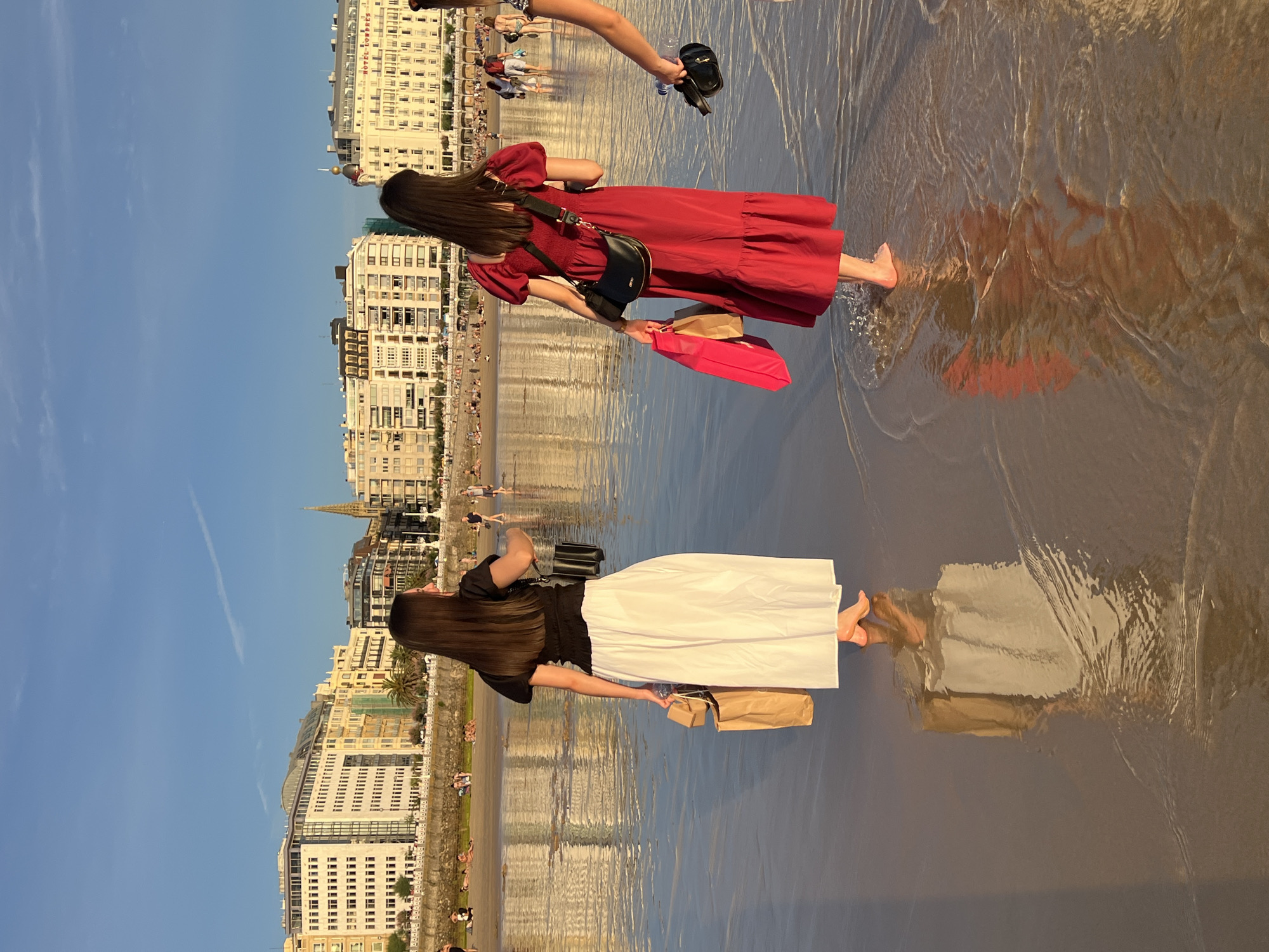 Two women walk along the shore of La Concha Beach in San Sebastián, Spain, near the promenade. The woman on the left wears a black top and a long white skirt, carrying a brown paper shopping bag and a smaller beige bag. The woman on the right wears a red dress and a black crossbody bag, holding a bright pink shopping bag and a brown paper shopping bag. Both women are barefoot, with shallow water lapping around their feet. In the background, the elegant buildings lining the promenade are visible, reflecting in the wet sand under a clear blue sky. Several other people are scattered on the beach and in the water, with more buildings and a church spire further in the distance. The scene is illuminated by the warm light of either a sunrise or sunset.