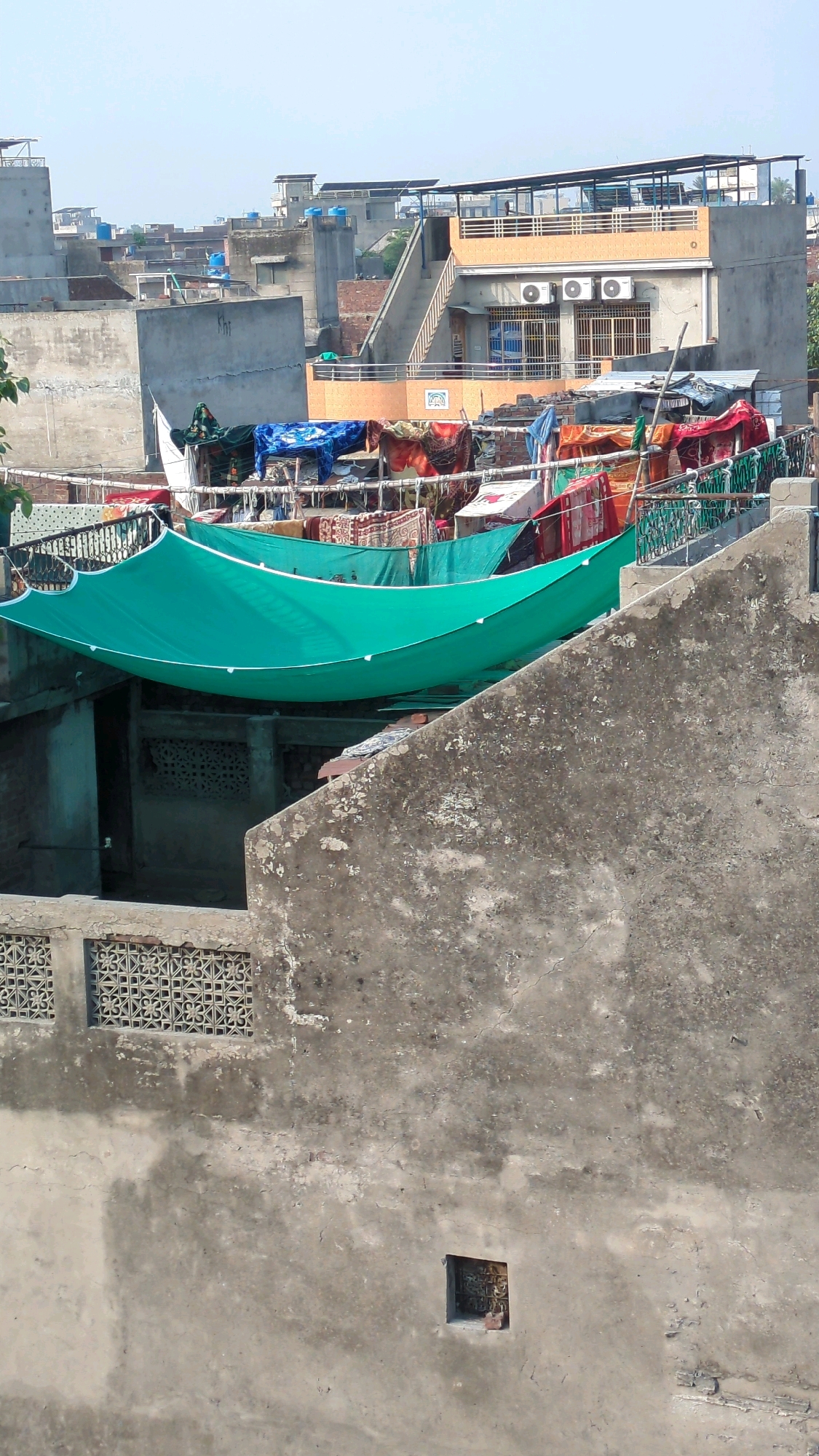 This image shows a view of rooftops in Lahore, Pakistan. In the foreground, a rough concrete wall dominates the lower portion of the frame, with a decorative perforated concrete block section visible on the left. Above this wall, a bright turquoise shade cloth is stretched taut, casting a shadow below. Behind the shade cloth, a jumble of items are visible on what appears to be a drying rack or clothesline, including colorful fabrics and blankets. In the mid-ground, a multi-story building with an orange-colored facade and blue accents is prominent. This building features an outdoor staircase leading to an upper level with a covered patio. Two air conditioning units are mounted on the facade of this building. Various other rooftops, some with satellite dishes and water tanks, are visible in the background, extending into the distance under a hazy sky, characteristic of an urban environment. The overall scene depicts a typical residential area in Lahore with evidence of daily life, such as clothes being dried outdoors.