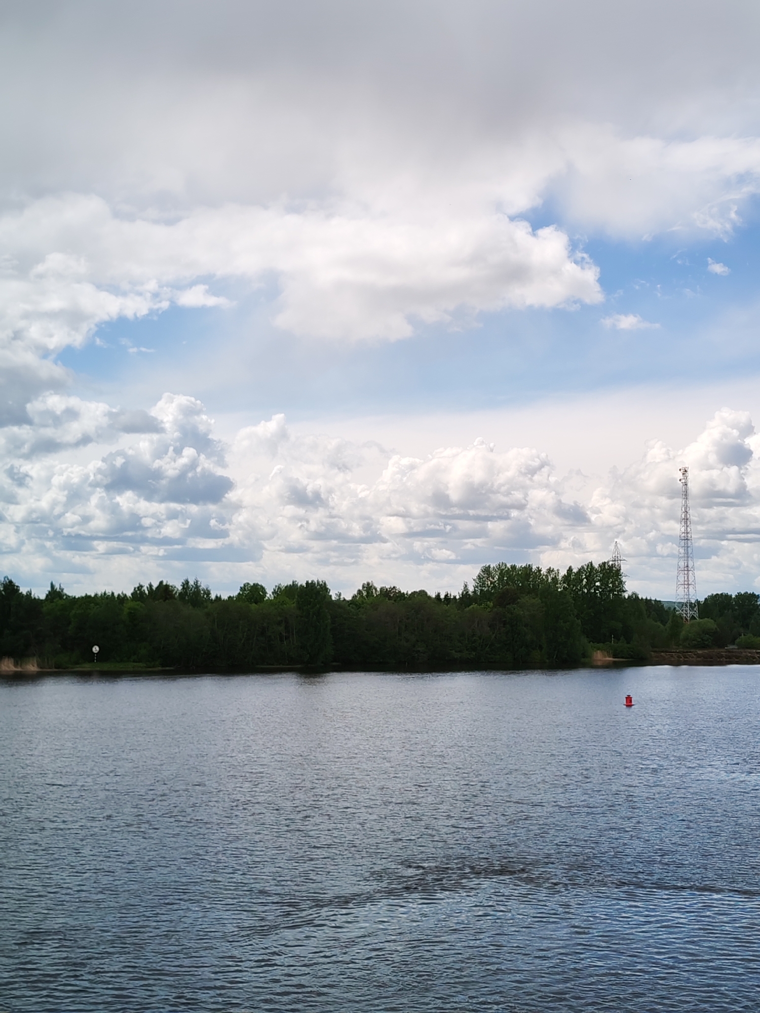 The image shows a body of water, likely a lake or wide river, under a cloudy sky. The sky features a mix of white, puffy cumulus clouds and patches of blue. In the distance, there is a treeline of green foliage along the opposite shore. A transmission tower is visible on the right, rising above the trees. There's a small red buoy in the water on the right side. The location is Белоусово, Russia.