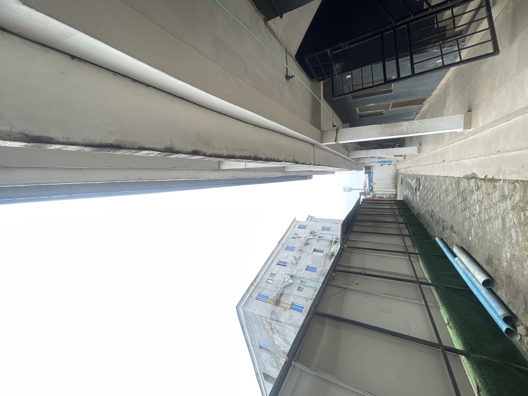 View of an urban alleyway in Ado, Nigeria. The shot is from a low angle, looking upwards between buildings. On the left, a tall, light-colored building extends into the sky. On the right, a narrower building with visible structural elements, including a metal-framed roof and white concrete supports, is present. Near the bottom right, lengths of blue PVC pipes rest on an artificial turf surface. Other details include clothes hanging on a line further down the alley and the overcast sky.
