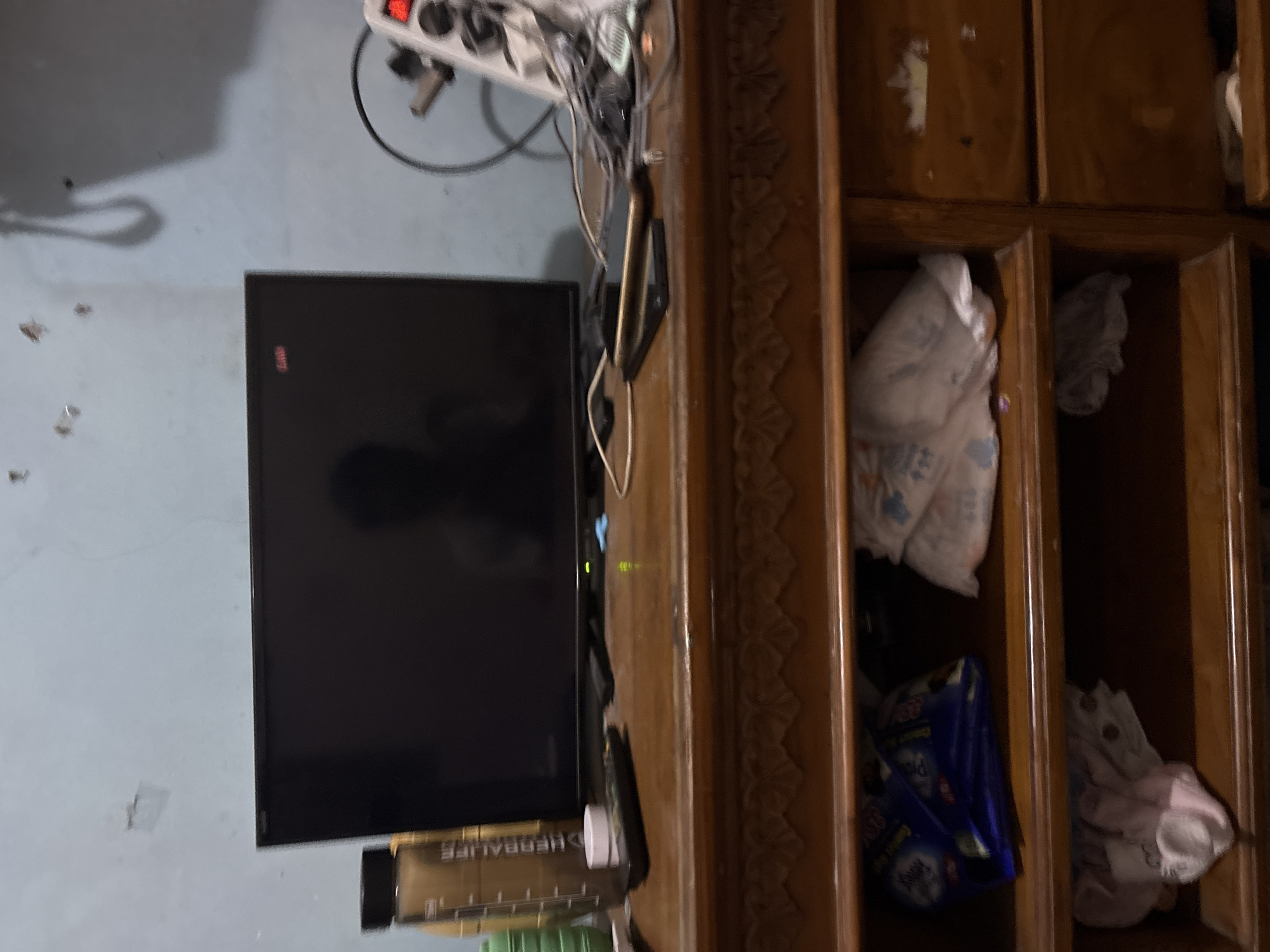 The image shows a wooden dresser with a television on top. The dresser has two shelves that are partially filled with various items, including a blue bag and white packages. The shelves appear to be in a residential setting, possibly a bedroom or living room. The wall behind the dresser is painted a light blue color. The dresser is located in the city of Palimanan, Indonesia.