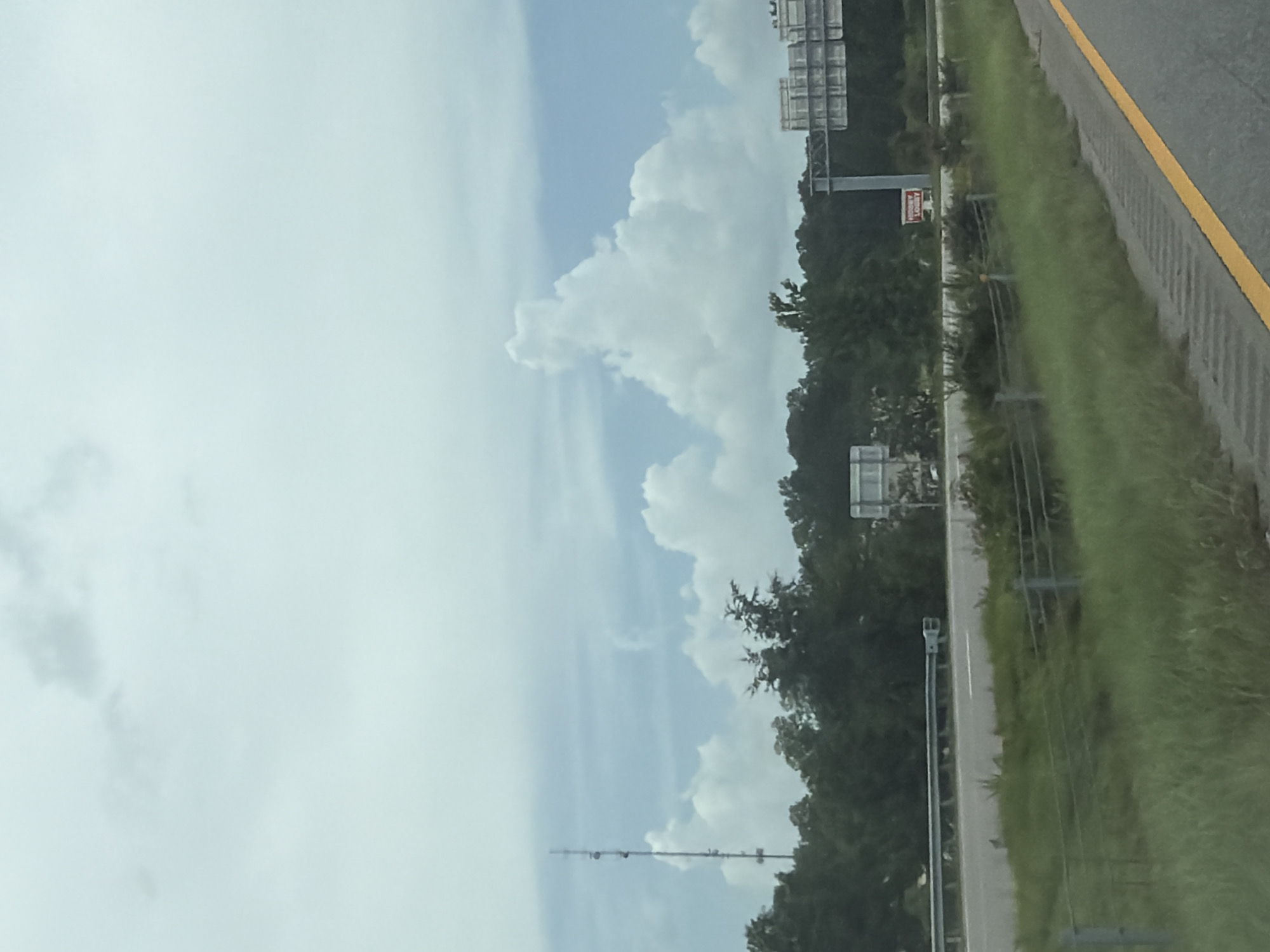 The image captures a cloudy sky over a highway scene, likely in Laurinburg, USA. In the distance, a tall radio tower stands against the sky. A line of lush green trees borders the highway, and on the right side of the road, a sign with red lettering is visible, reading "HOBBY LOBBY". The highway itself is paved, with a yellow line marking the edge of the road. The foreground shows a grassy verge, partially obscured by a wire fence. The overall scene is one of a typical roadside view on an overcast day.