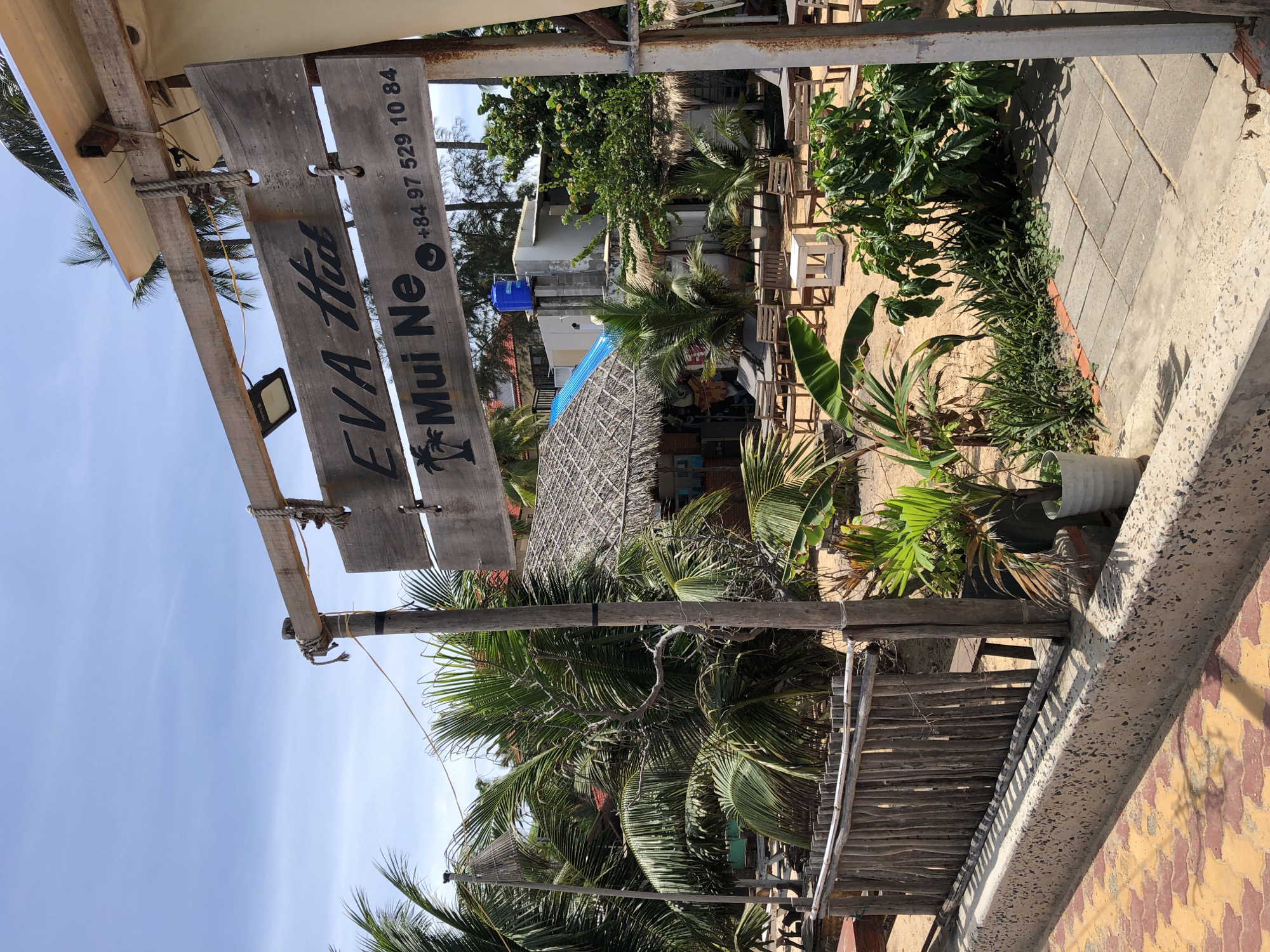 The image shows a wooden sign for "EVA Hut Mui Ne" with a phone number "+84 97 529 10 84" and a palm tree silhouette. The sign is hanging from wooden posts and ropes, with a light fixture attached to the top. Behind the sign, there are palm trees, thatched-roof huts, and beach chairs set up on a sandy area. In the foreground, there is a paved walkway and a low wooden fence. The sky is blue with some clouds. The overall scene suggests a beach resort or restaurant in Mũi Né, Vietnam.