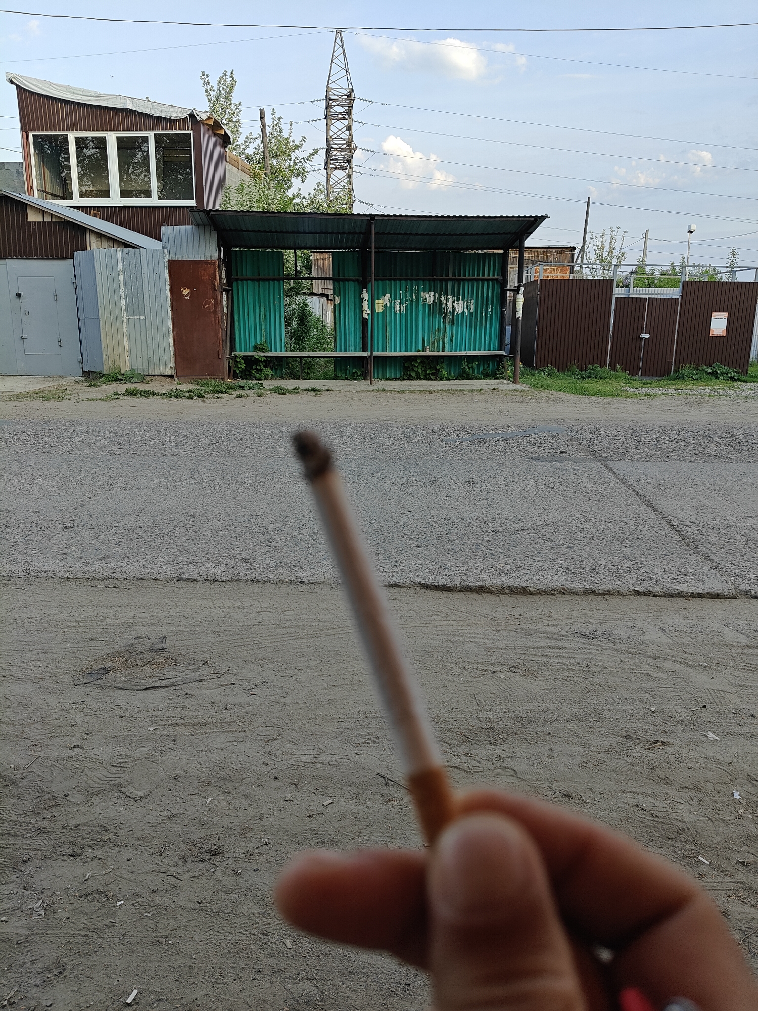 The image depicts a hand holding a lit cigarette in the foreground, partially obscuring the view of a street scene. In the background, a simple bus stop with green corrugated metal walls and a dark metal roof is visible. To the left of the bus stop is a gray metal structure, possibly a garage or storage unit, with a similar structure further to the left. A brown metal fence borders the right side of the bus stop.
A street or road stretches across the midground. Beyond the road, a power line tower rises in the distance. The sky is overcast, with visible power lines. The surrounding area appears somewhat industrial or semi-rural. Given the stated location, this scene is likely in or around the city of Chaikovsky, Russia.
