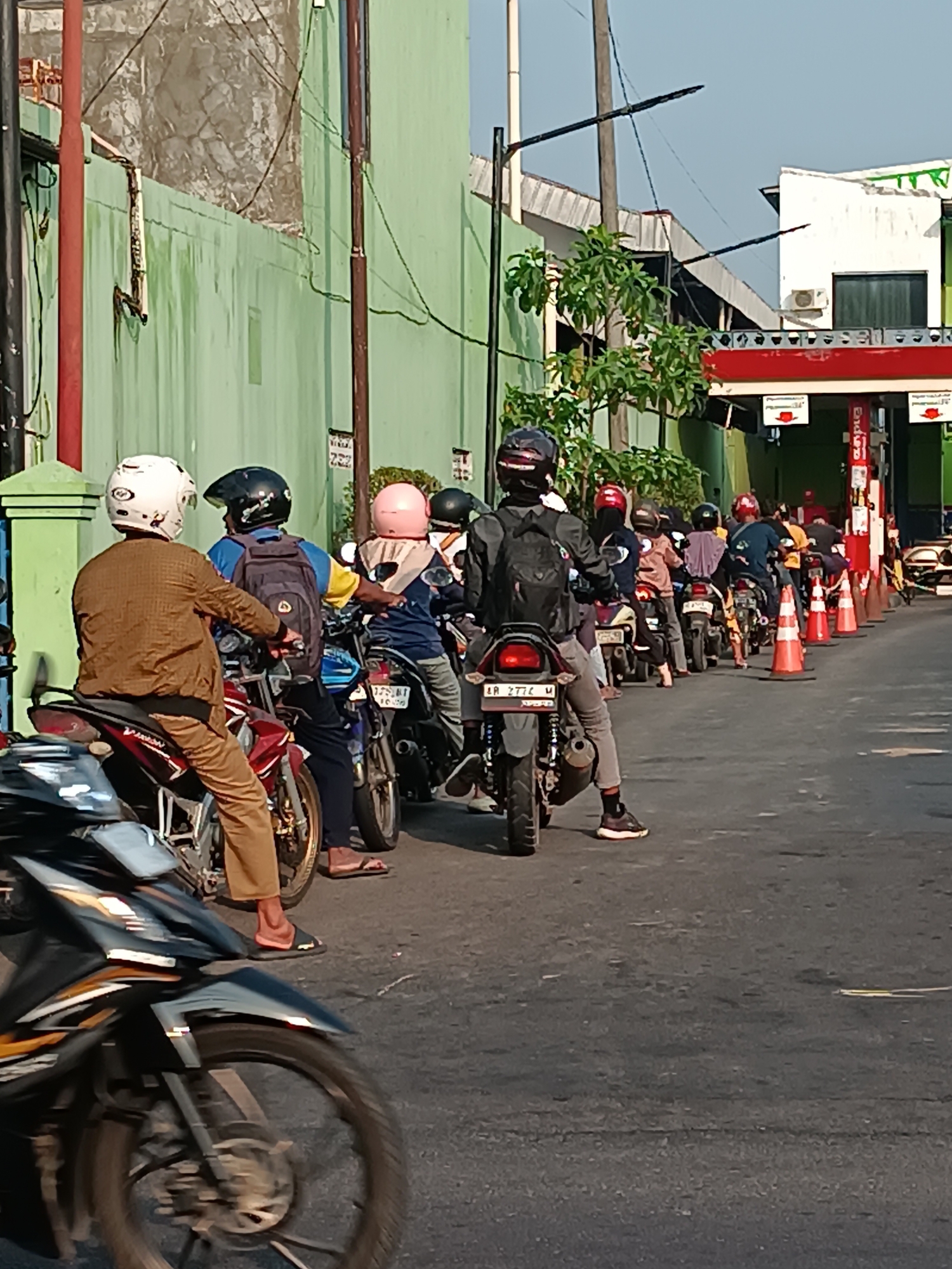 The image depicts a queue of motorcycles and their riders at a gas station in Imogiri, Indonesia. The gas station has a green building and a red awning. Several people are waiting on their motorcycles, some wearing helmets. The ground is asphalt. The weather appears to be sunny.